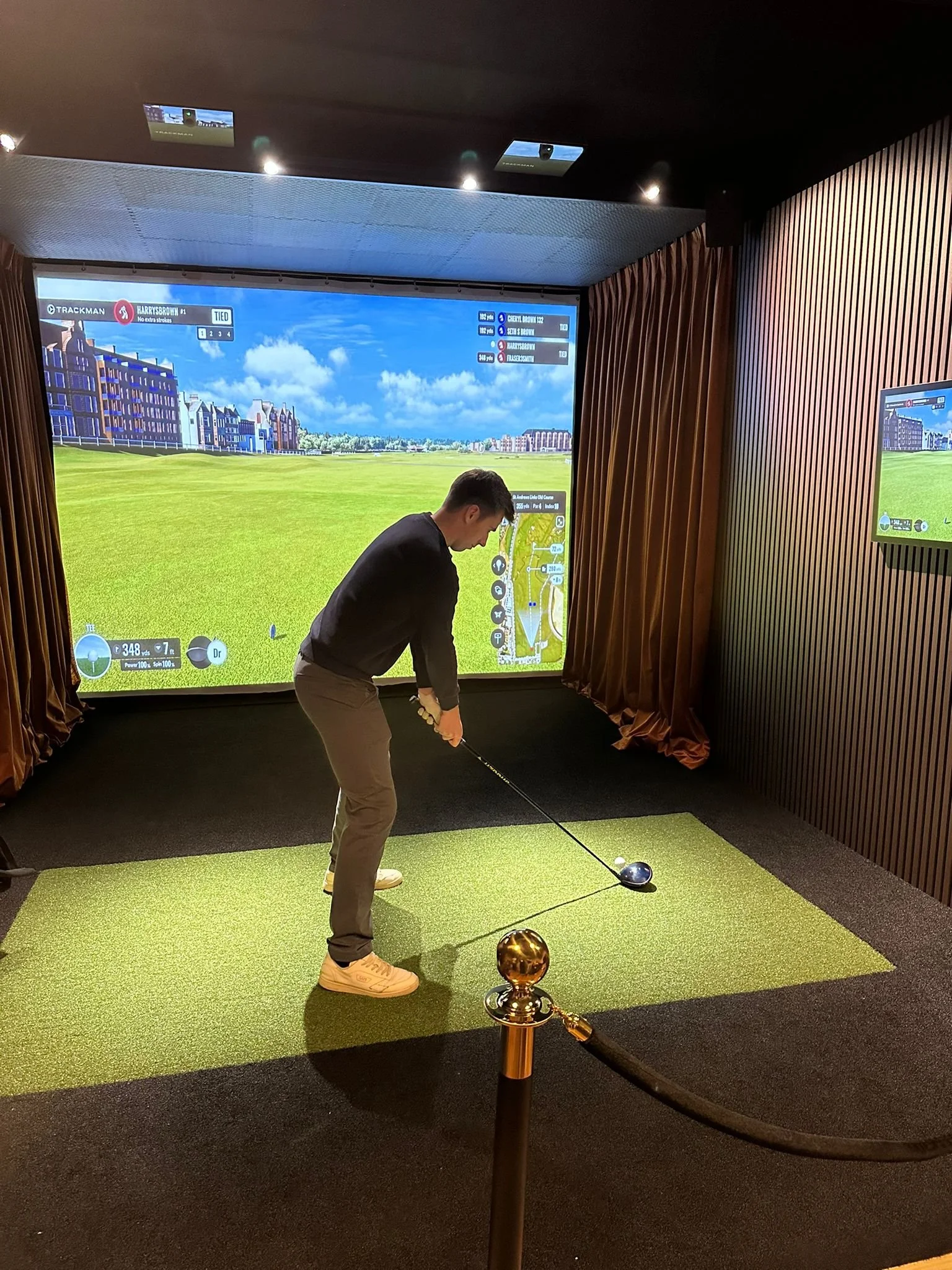 Man playing virtual golf in indoor golf simulator, with large screen displaying golf course.