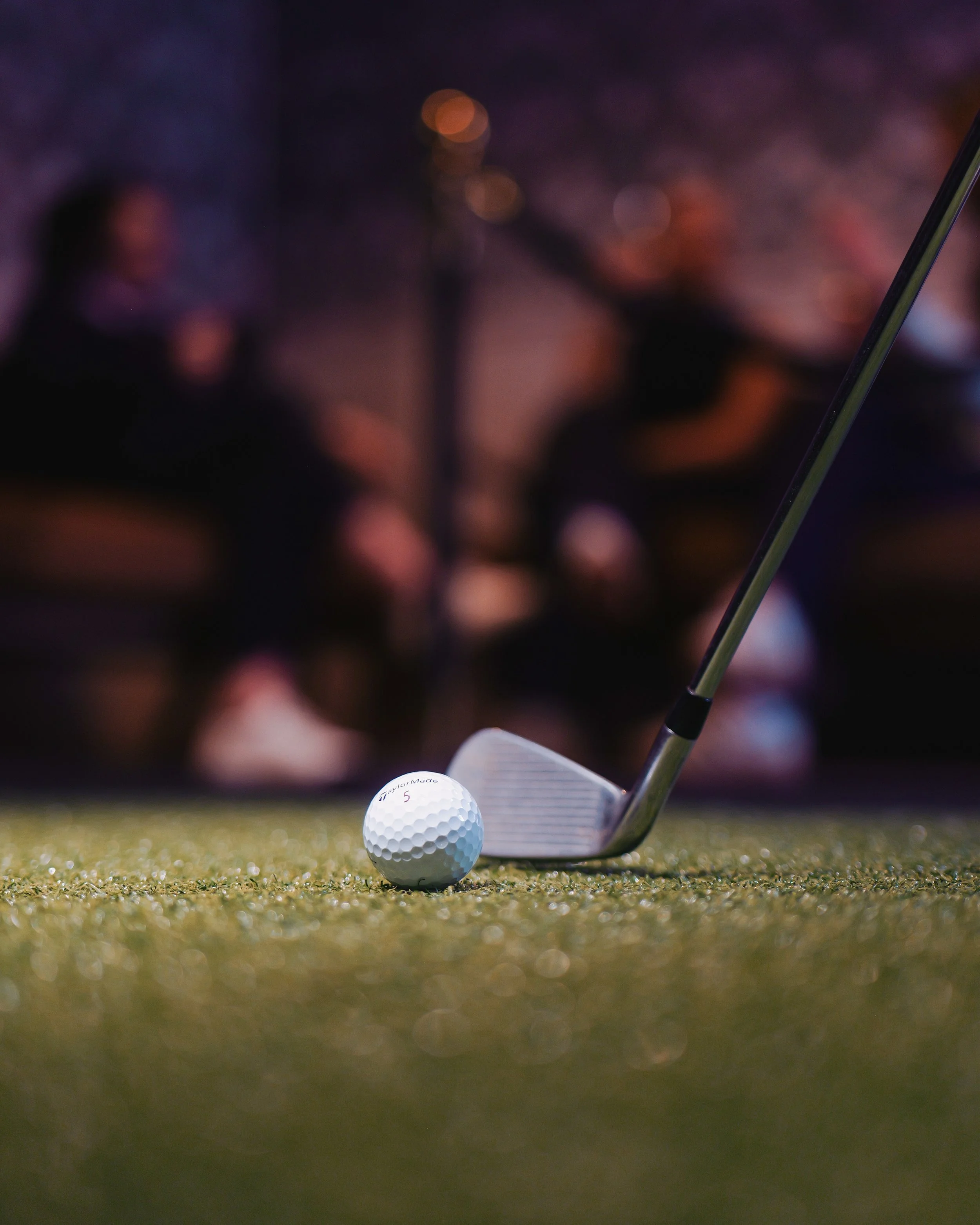 A golf ball on a putting green with a golf club behind it, and two blurred people sitting in the background under string lights.