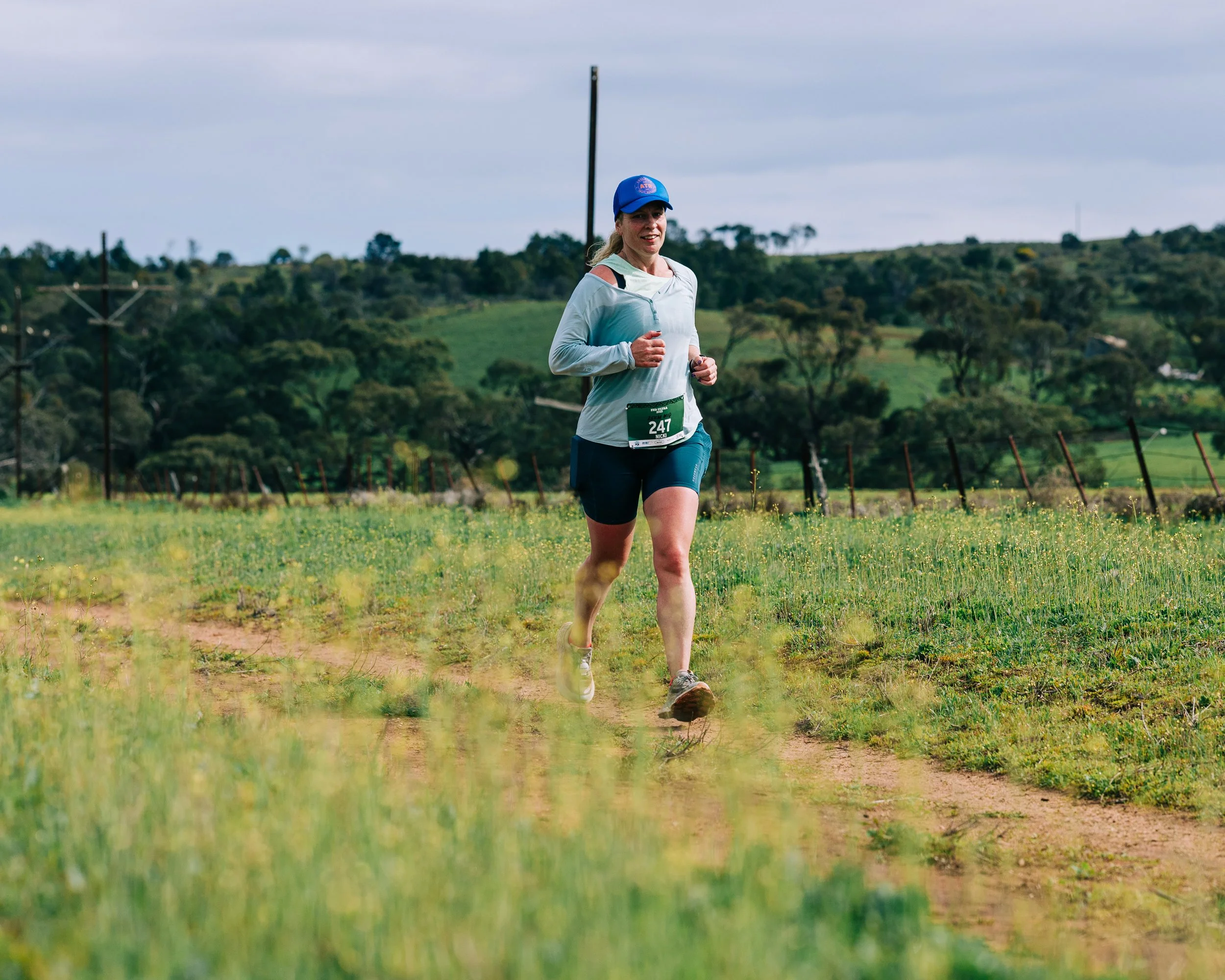 Female runner wearing a blue cap and athletic clothing running outdoors on a dirt trail in a rural area with green hills and trees in the background.