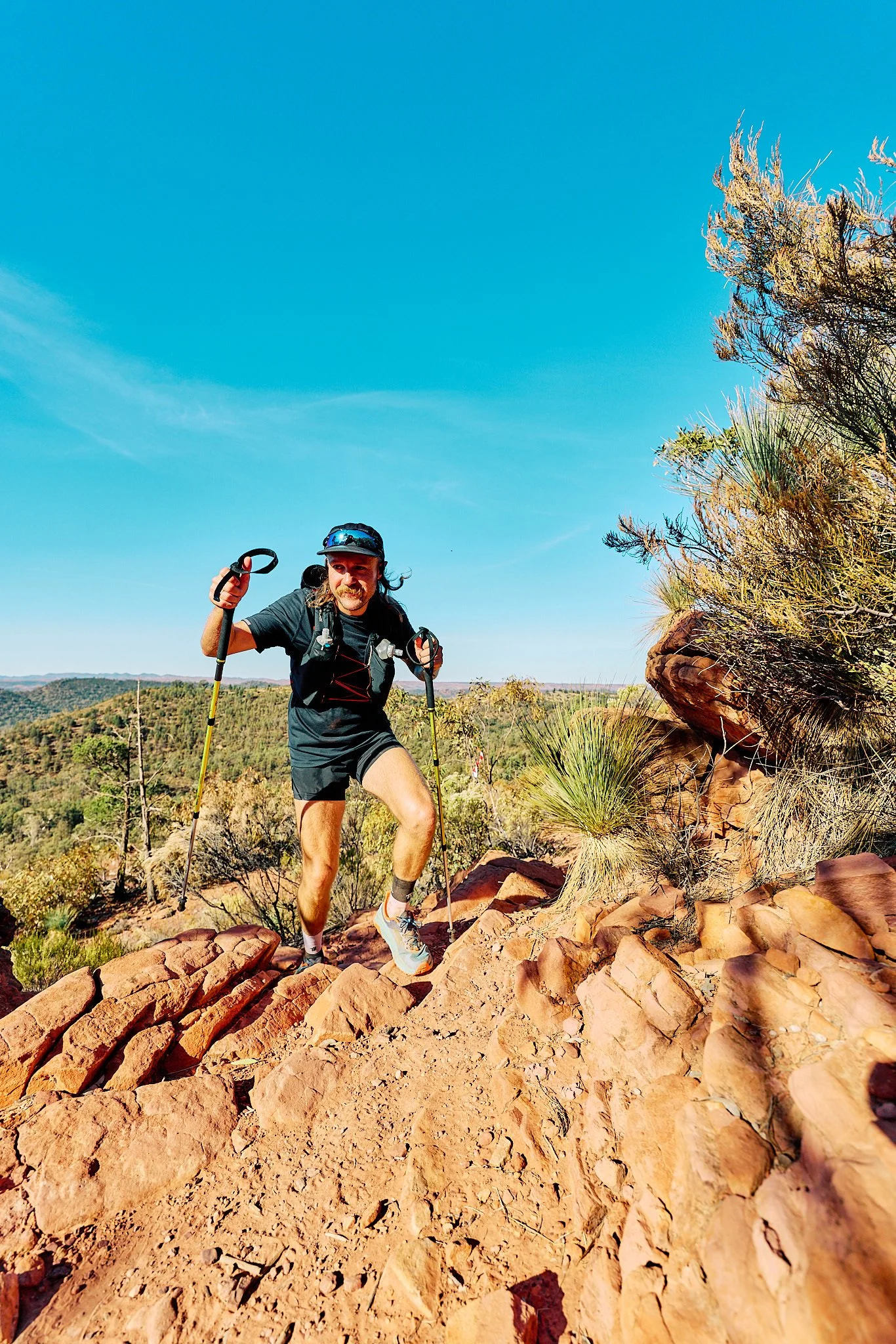 Four young adults running and celebrating on a trail in a grassy area with trees, wearing athletic clothes and race bibs.