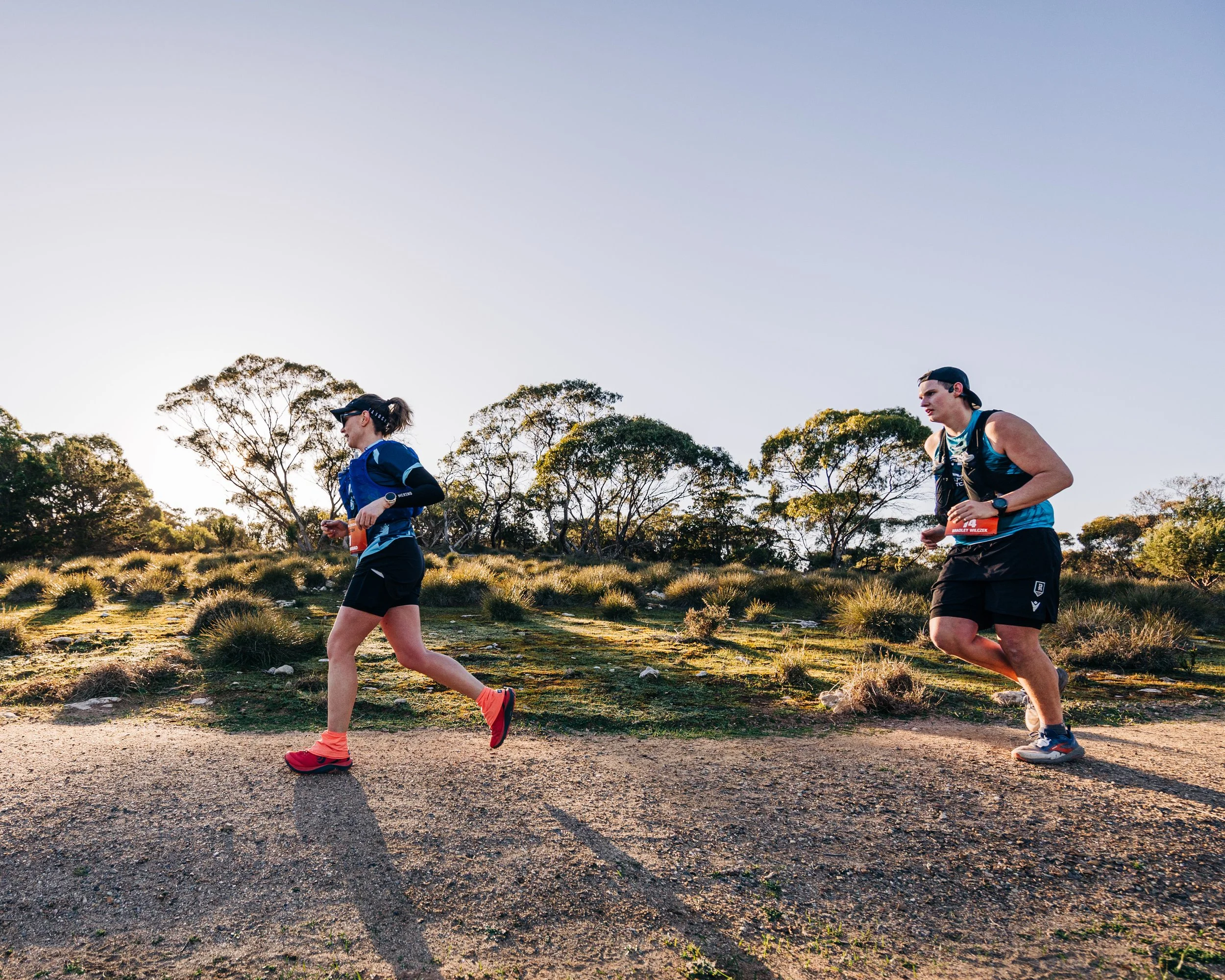 Two runners, a woman and a man, running on a trail in a scenic outdoor area with trees and shrubs, during what appears to be late afternoon or early evening sunlight.