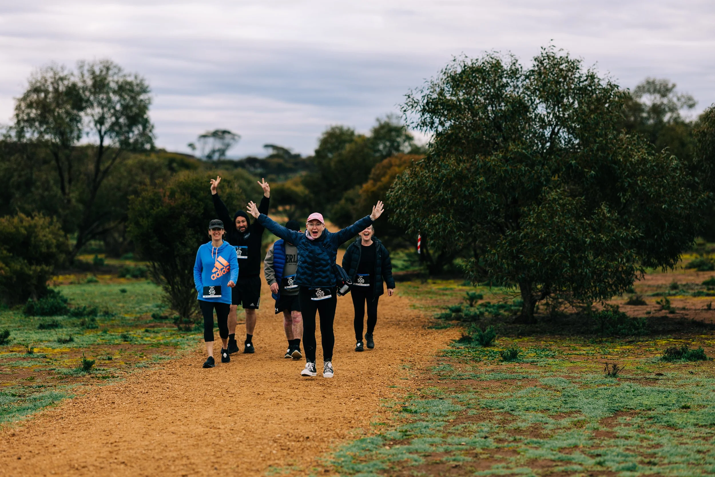 Group of five people running or walking outdoors on a dirt trail, smiling and raising their arms, in a natural setting with trees and cloudy sky.