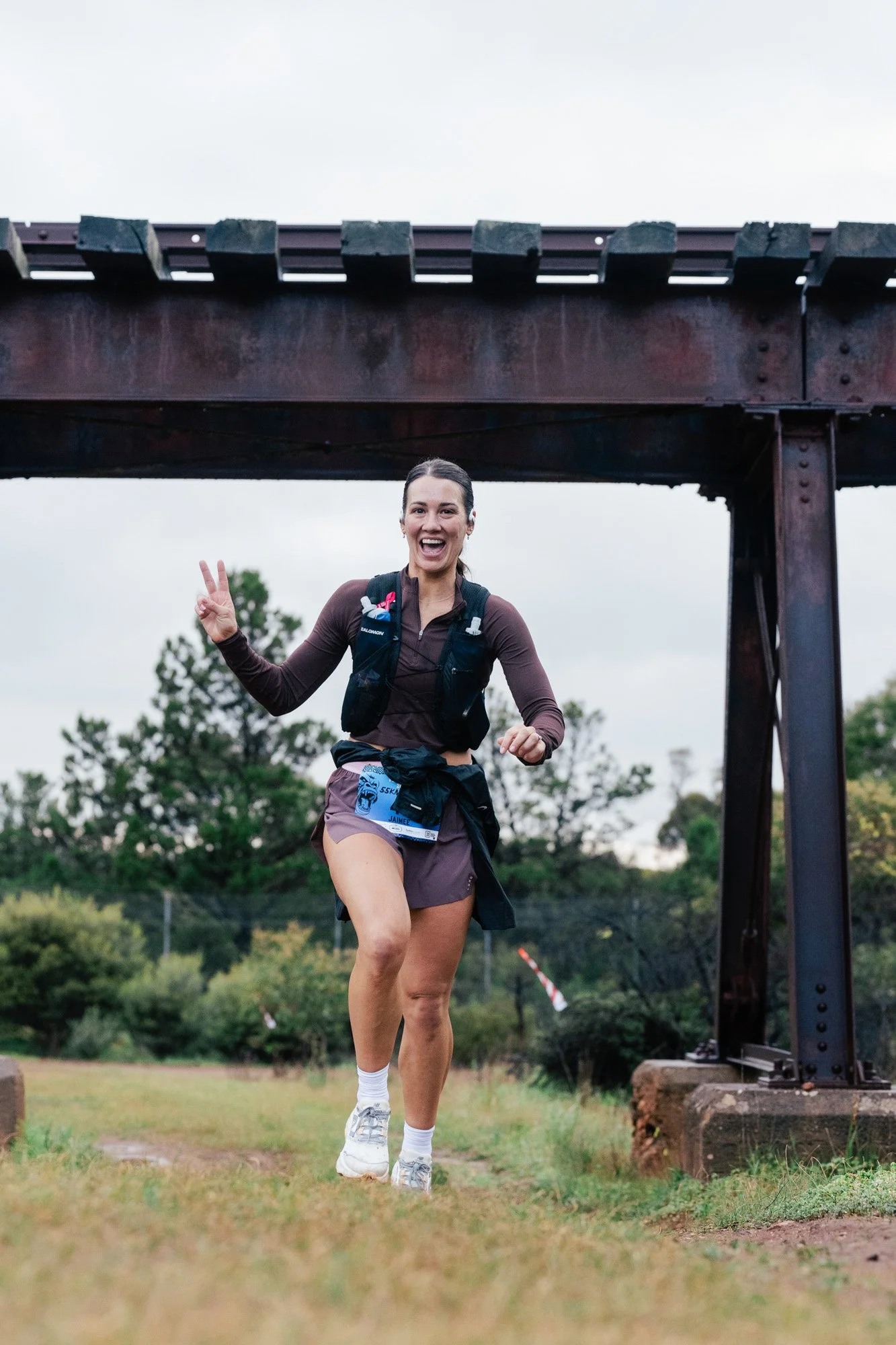 A person wearing a yellow jacket, black shorts, and a black hat running on a dirt trail in a mountainous area with greenery and blue sky.