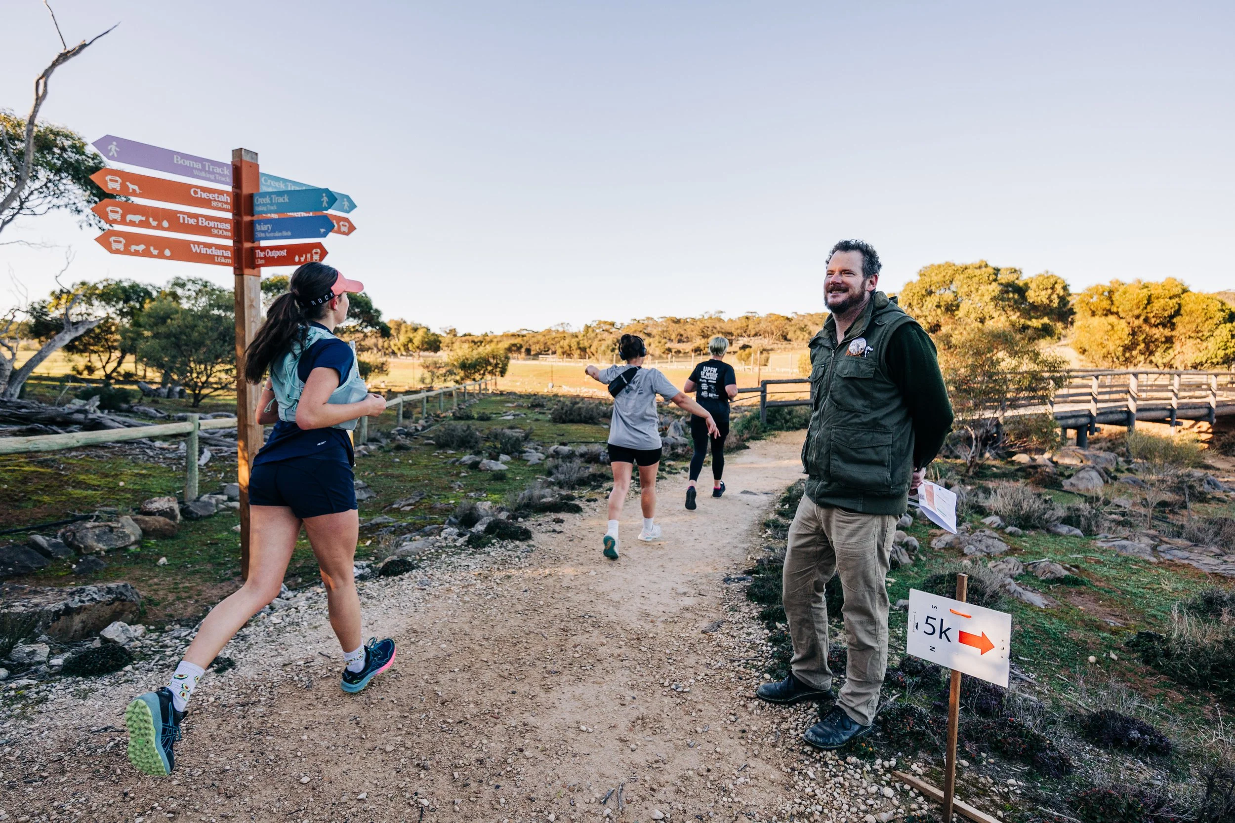 People running on a trail at a park with directional signs and a man standing by a 5K sign.