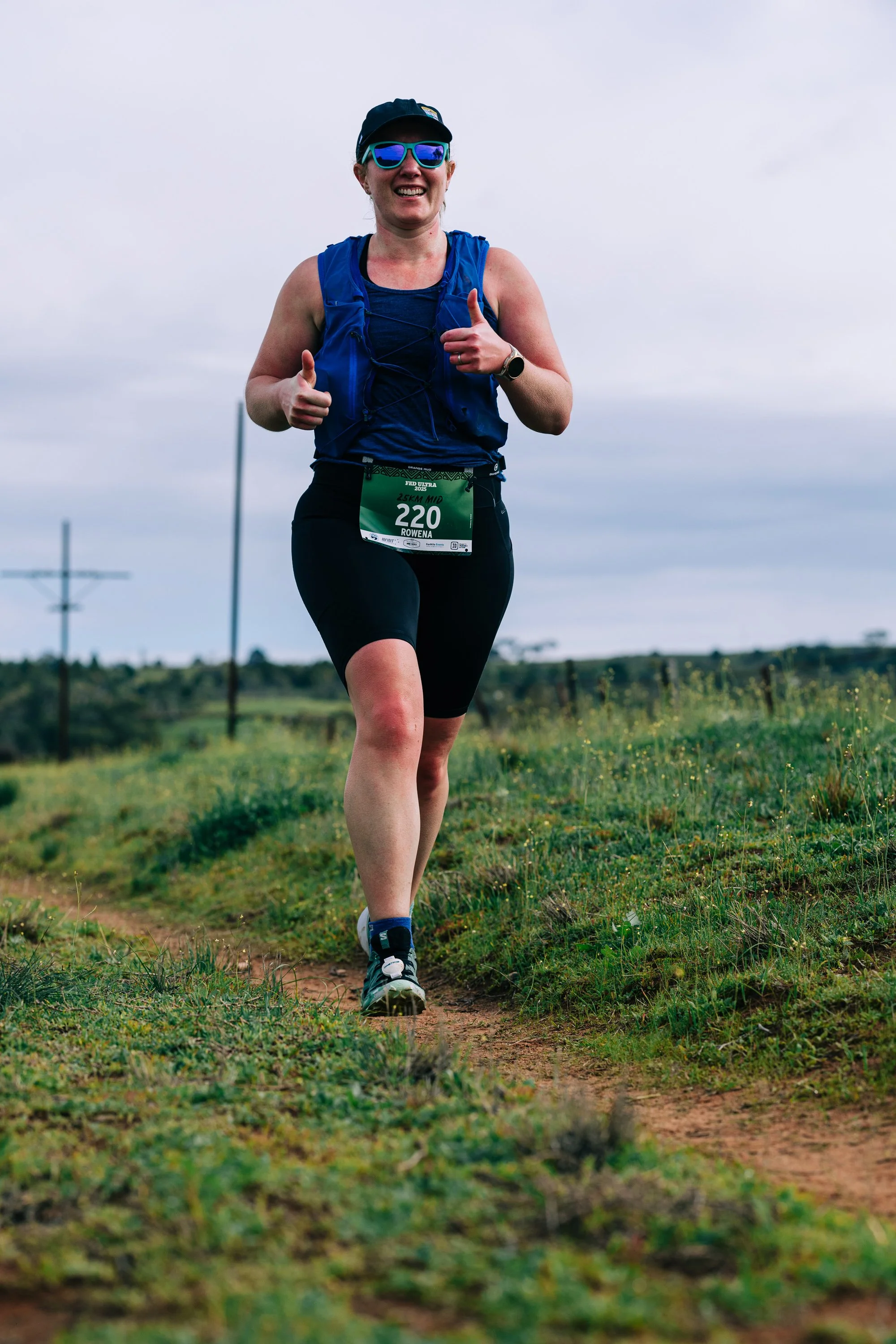 A woman running outdoors on a dirt trail, smiling, wearing sunglasses, a blue vest, black athletic pants, and a running bib numbered 220.