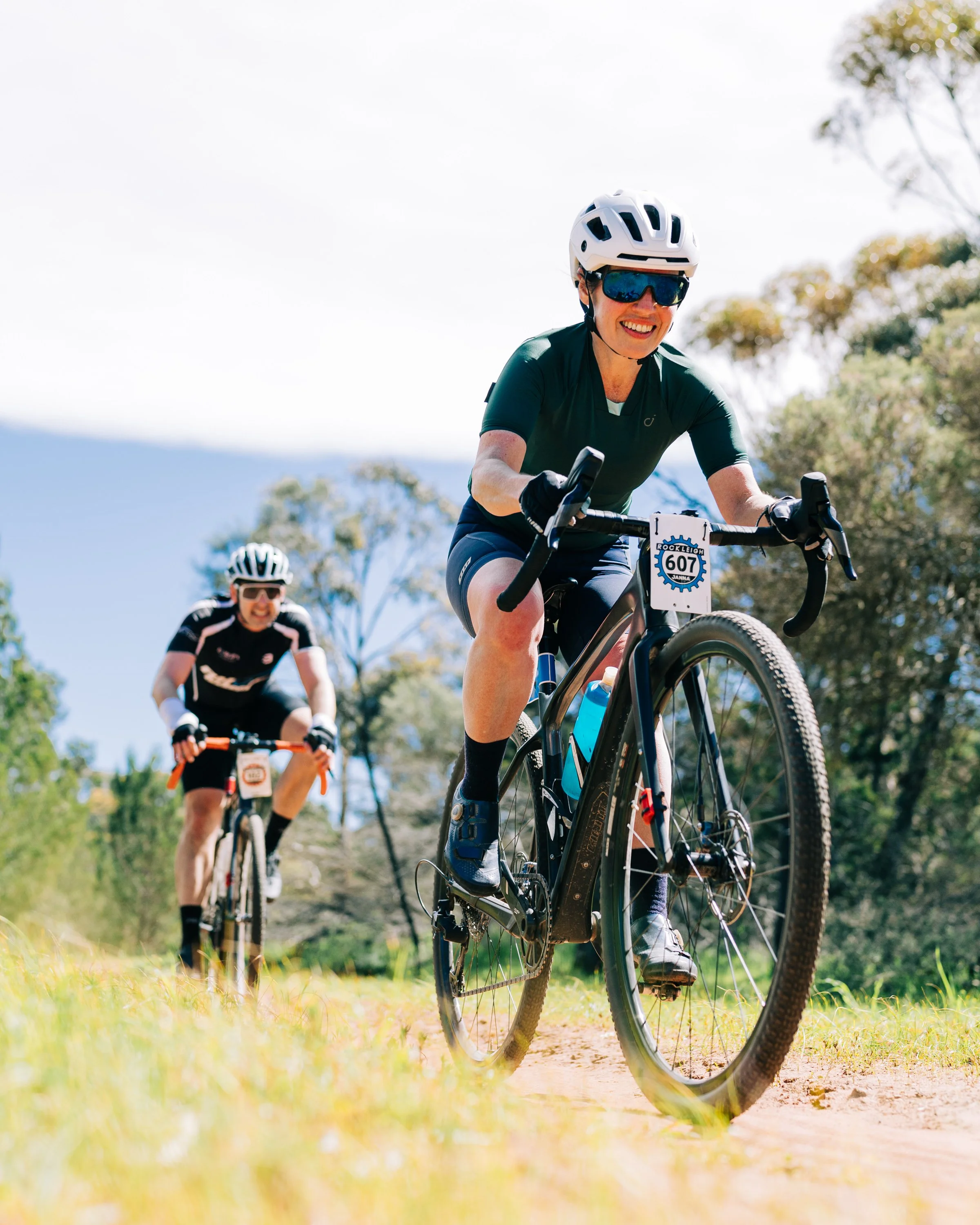 Four cyclists riding on a dirt path through green fields under a partly cloudy sky.