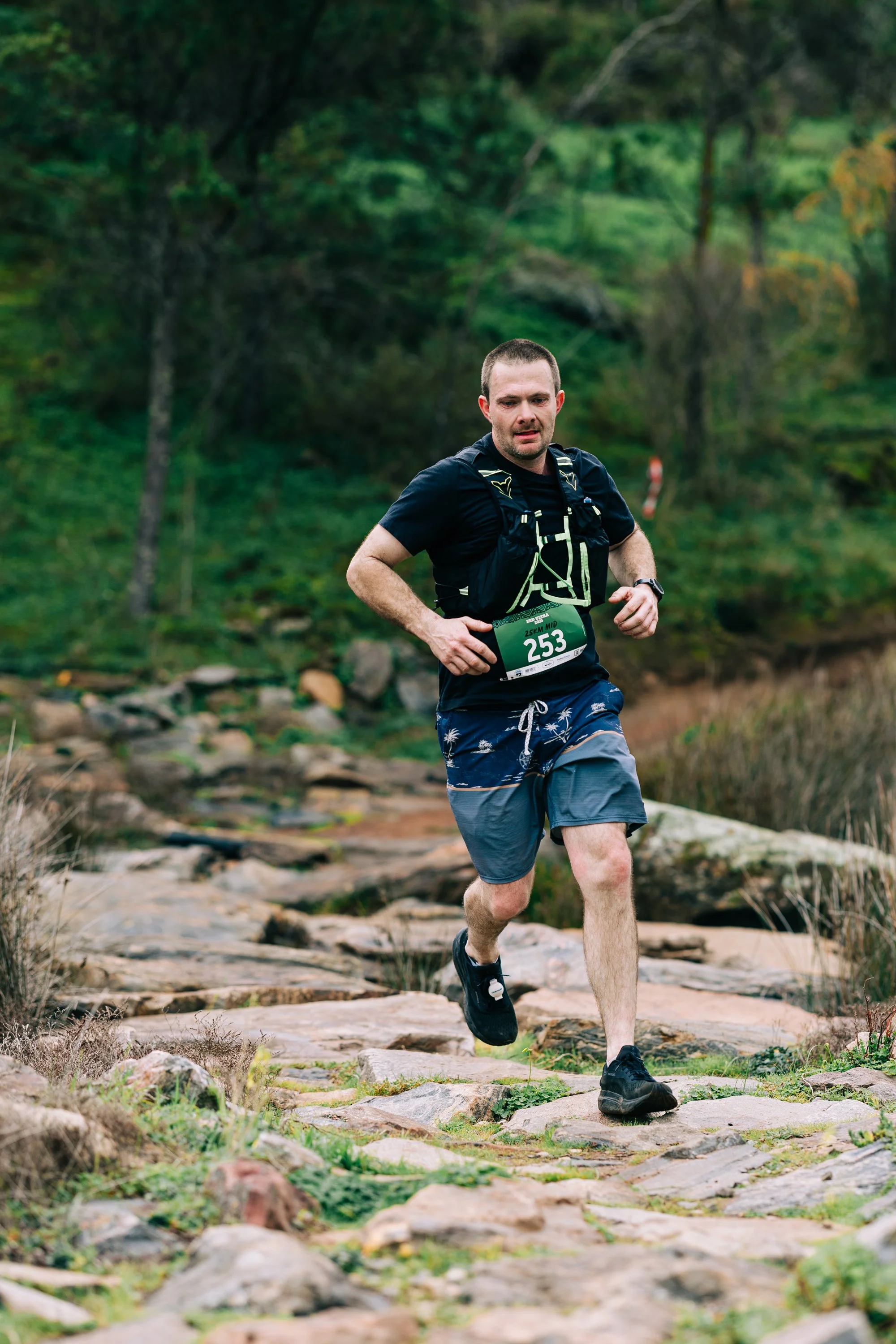 A man running on a rocky trail in a forest during a race, wearing a bib number 253, black shirt, and blue shorts.