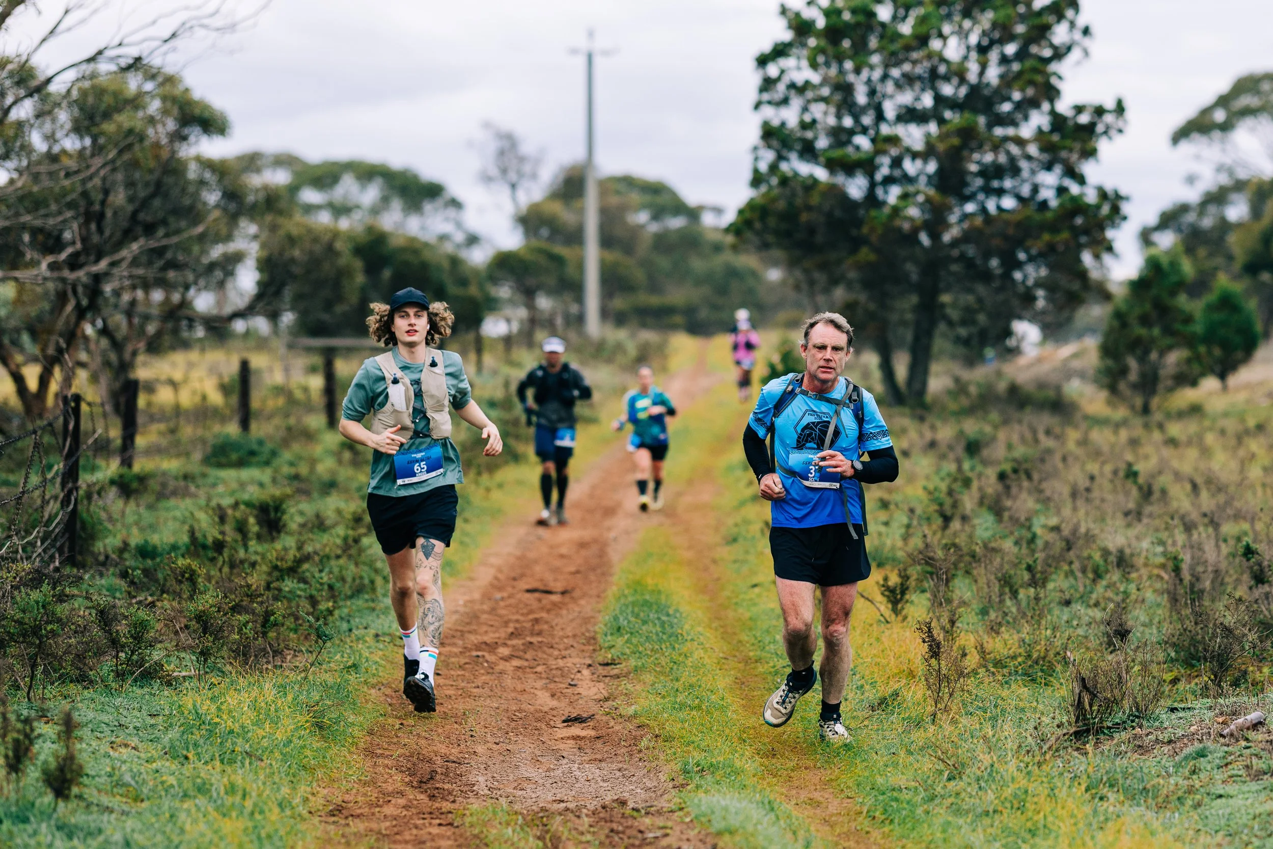 Group of runners participating in a trail race on a dirt path through a green, wooded area with overcast sky