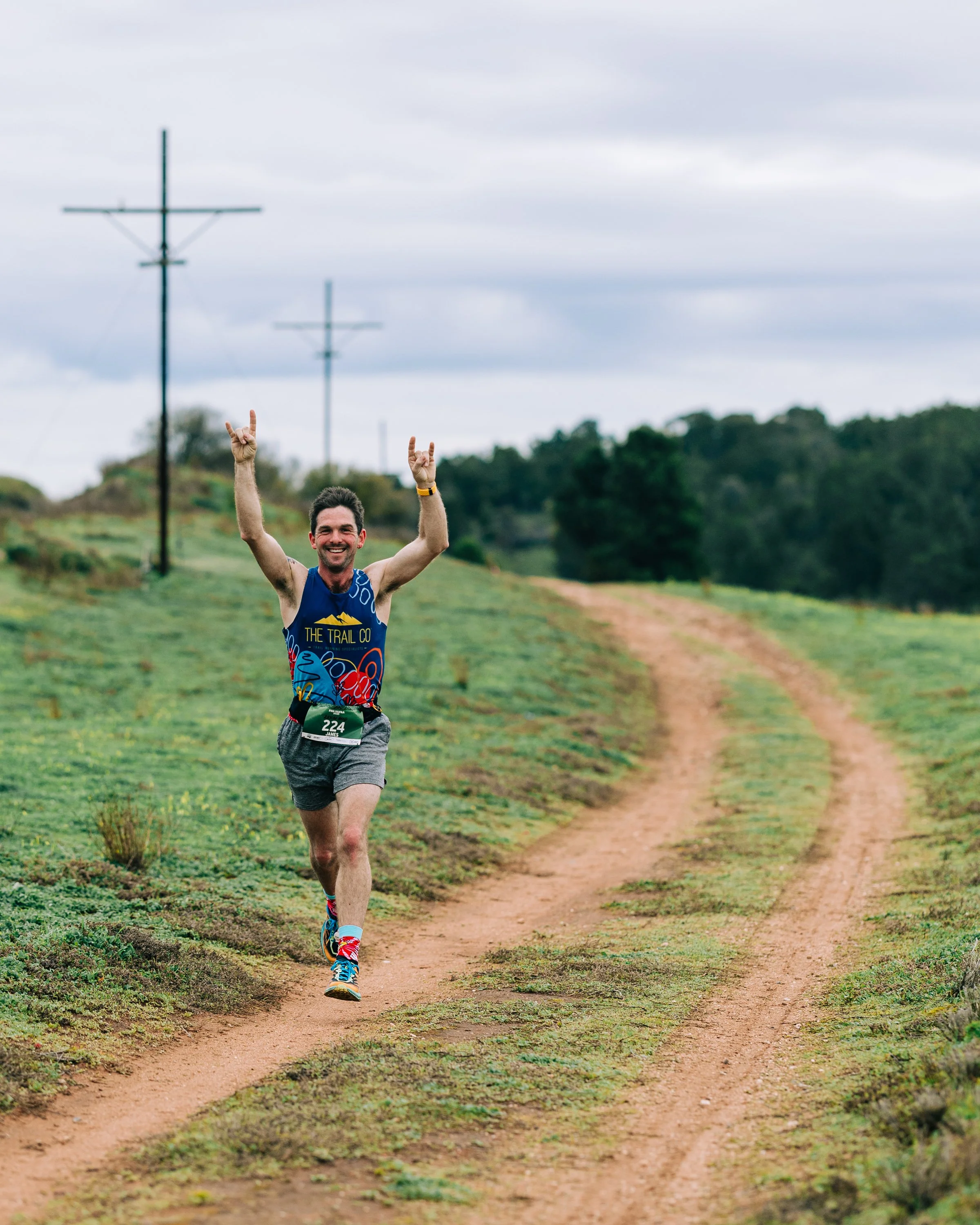 Man running on a dirt trail with his arms raised in celebration, smiling, surrounded by green grassy fields and trees, with power lines in the background.