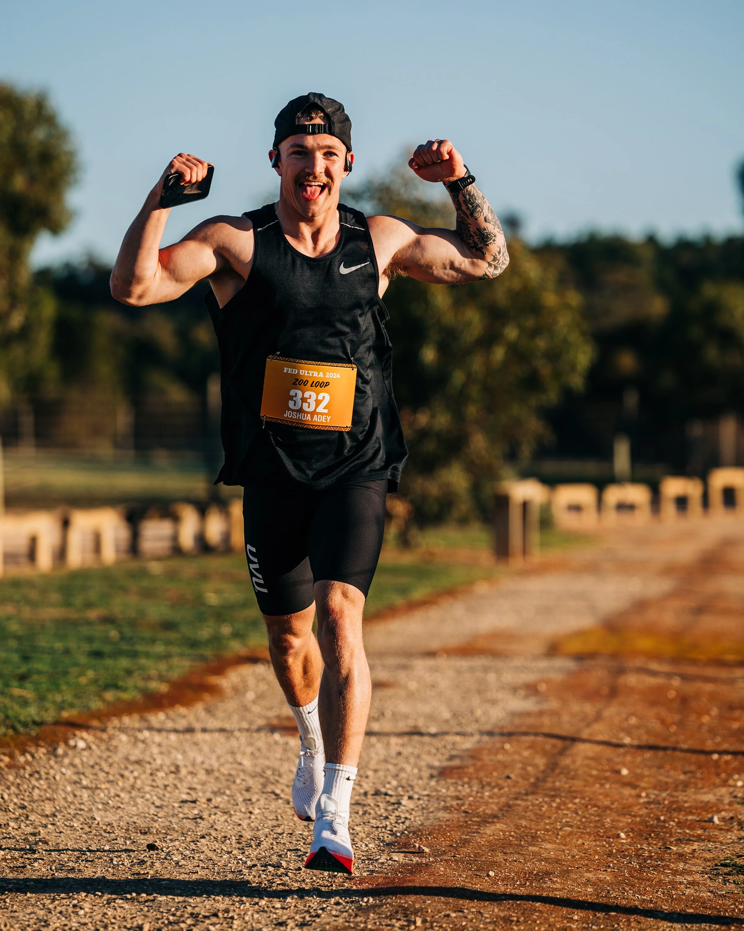 Man running on a gravel trail during a race, raising his arms in celebration, wearing a black sports outfit, a cap backward, and a race bib numbered 332.