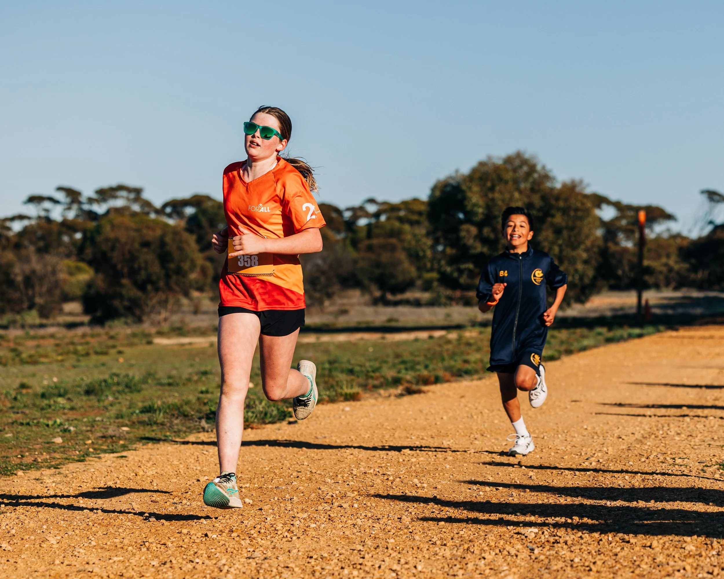 Two children running on a dirt track outdoors on a sunny day.