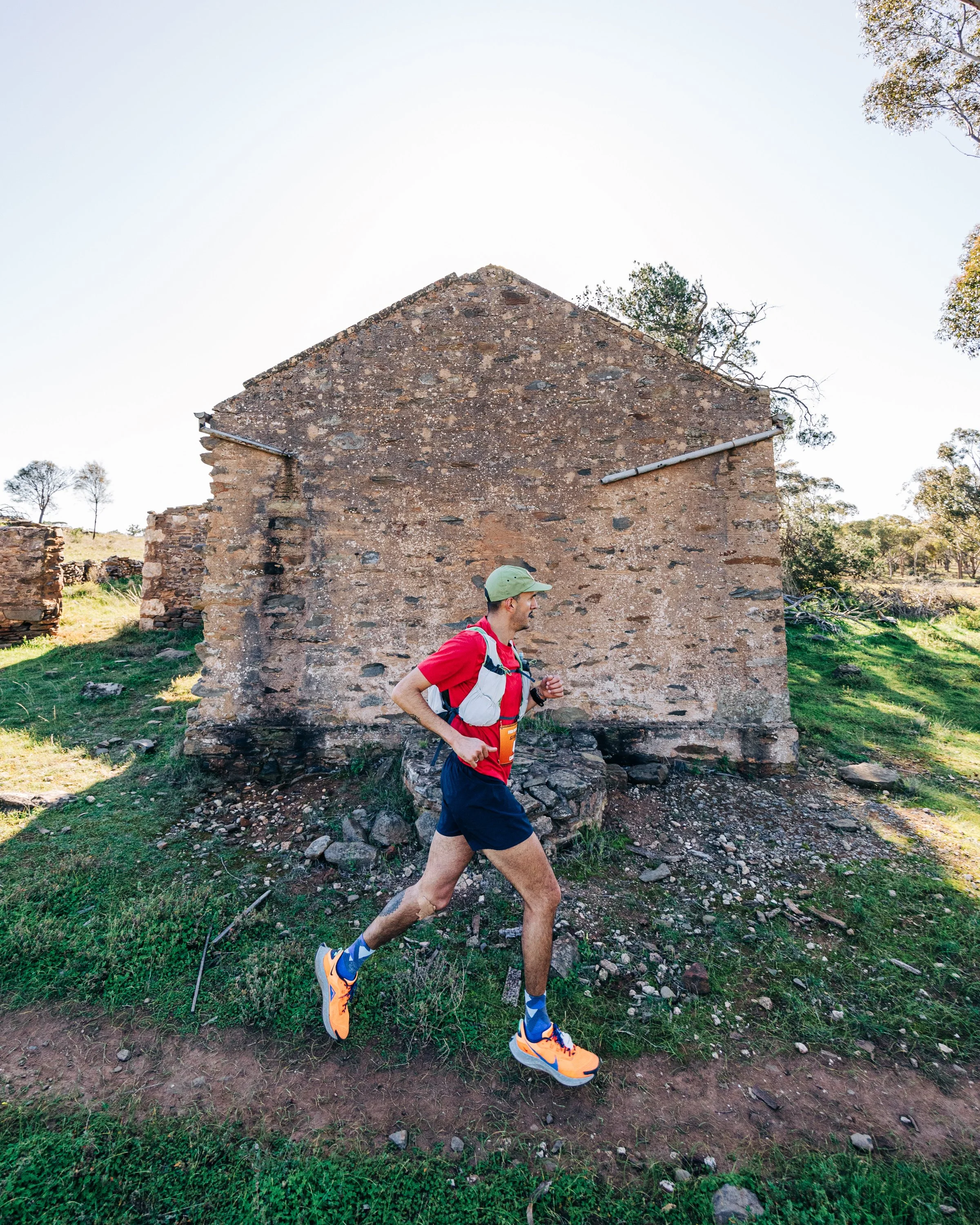 A man running outdoors in athletic gear including an orange and blue running shoe, a red shirt, black shorts, a green cap, and a hydration pack, on a dirt trail in front of an old stone building with damaged walls and a pitched roof, surrounded by gr