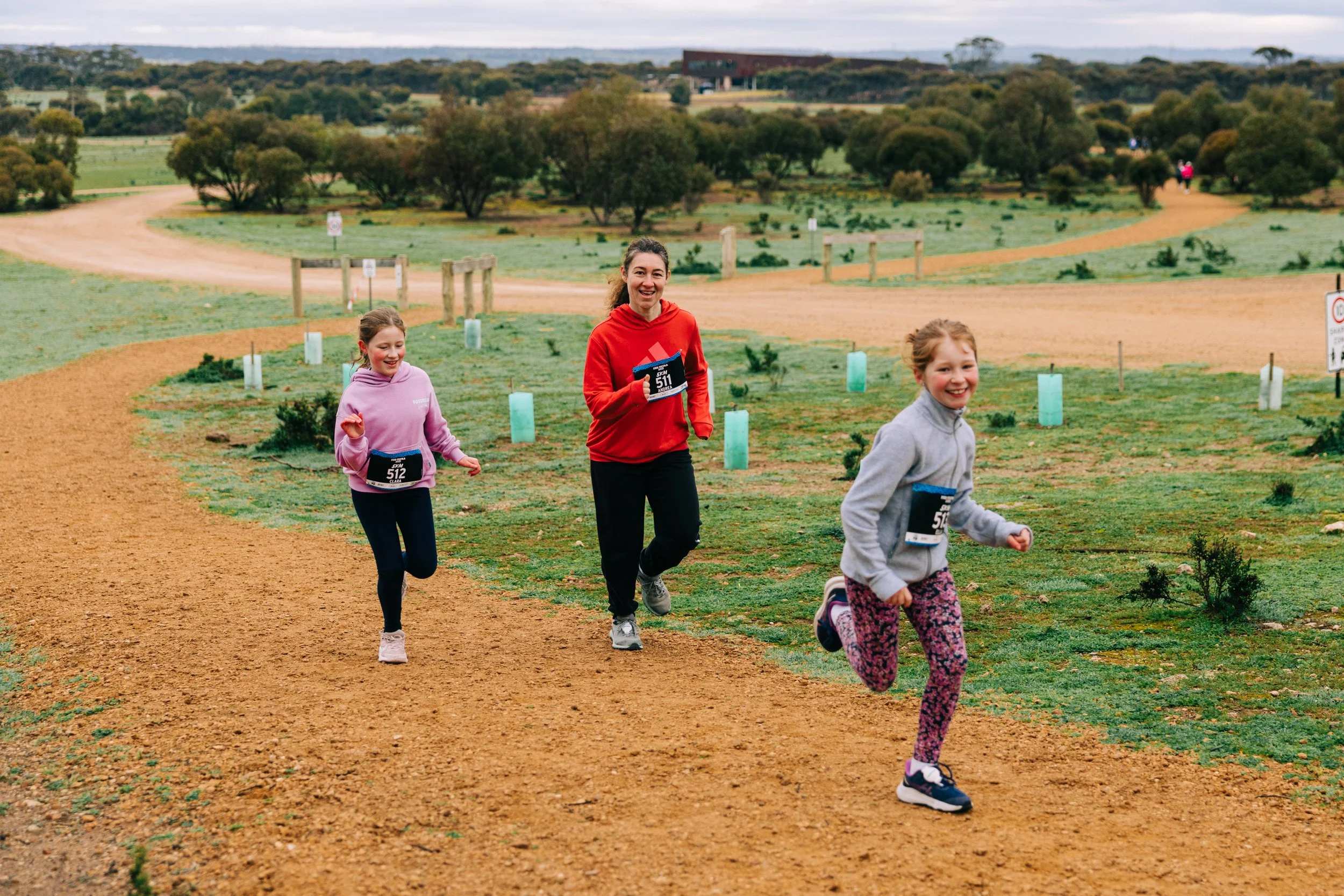 Three females running on a dirt trail through a park with greenery and trees, overcast sky, all wearing athletic clothing and race bibs.