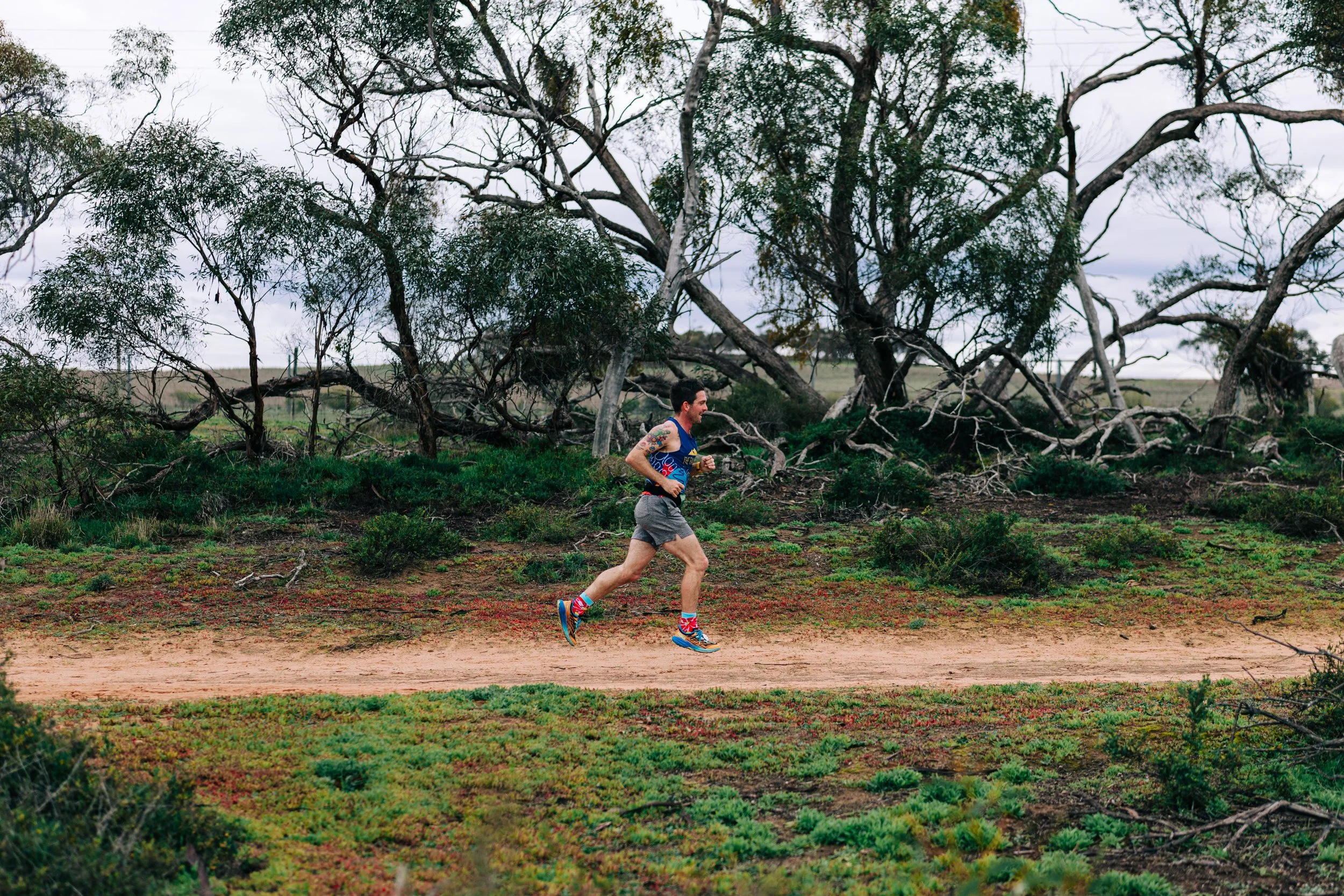 A man runs on a dirt trail in a natural landscape with scattered green shrubs and leafless trees under an overcast sky.