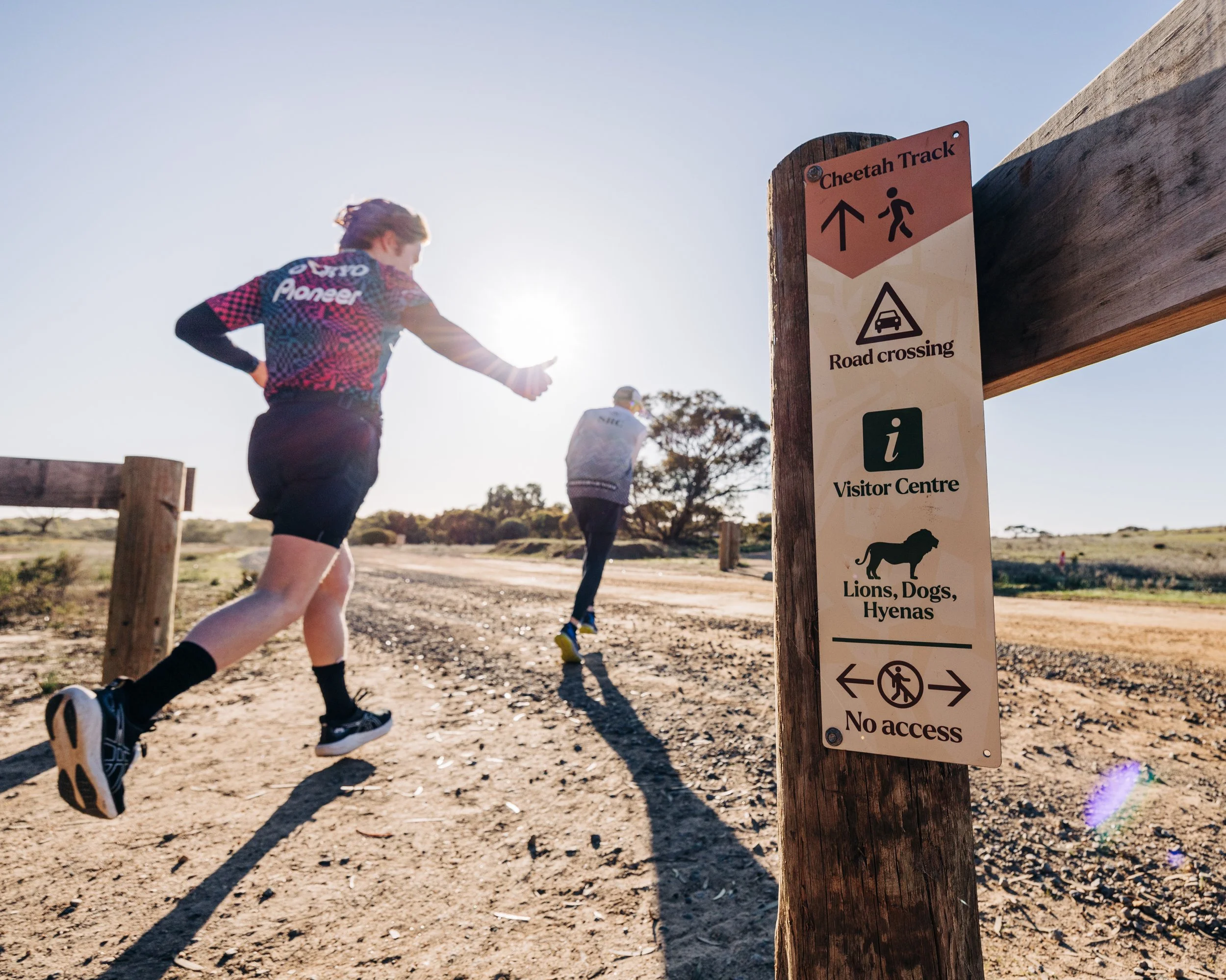 Two runners on a dirt trail in a natural park, with a sign in the foreground indicating a Cheetah Track, road crossing, visitor center, and no access for lions, dogs, and hyenas.