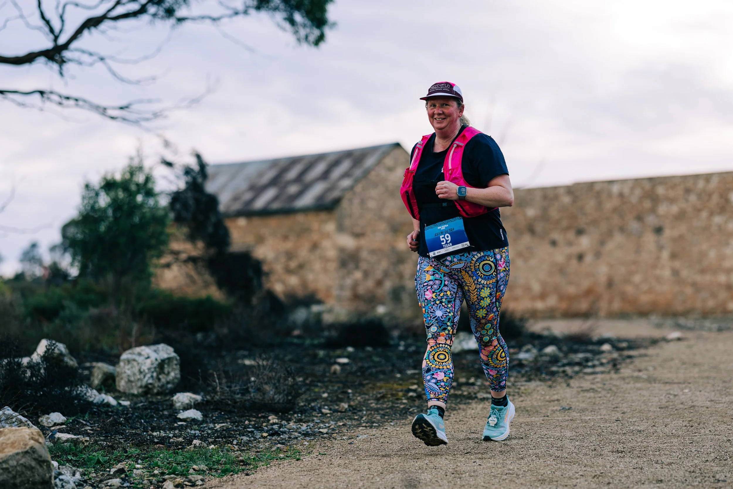 A woman running outdoors on a dirt trail with a stone building and some trees in the background, wearing colorful leggings, a black shirt, a pink vest, and a cap.
