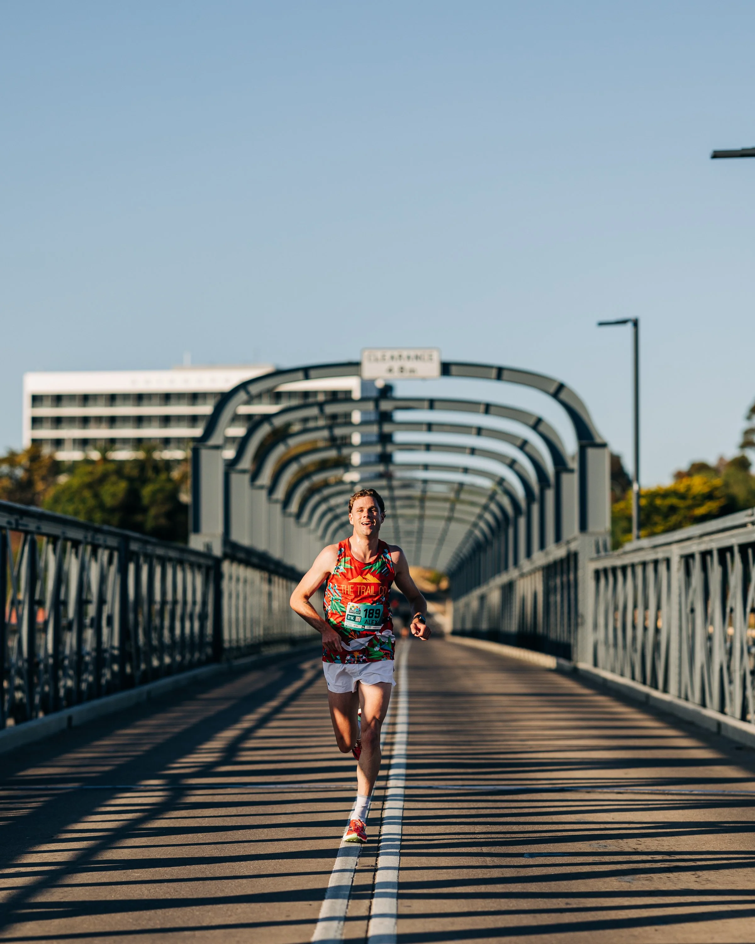 A male marathon runner in a colorful tank top and white shorts running on a bridge during daylight.