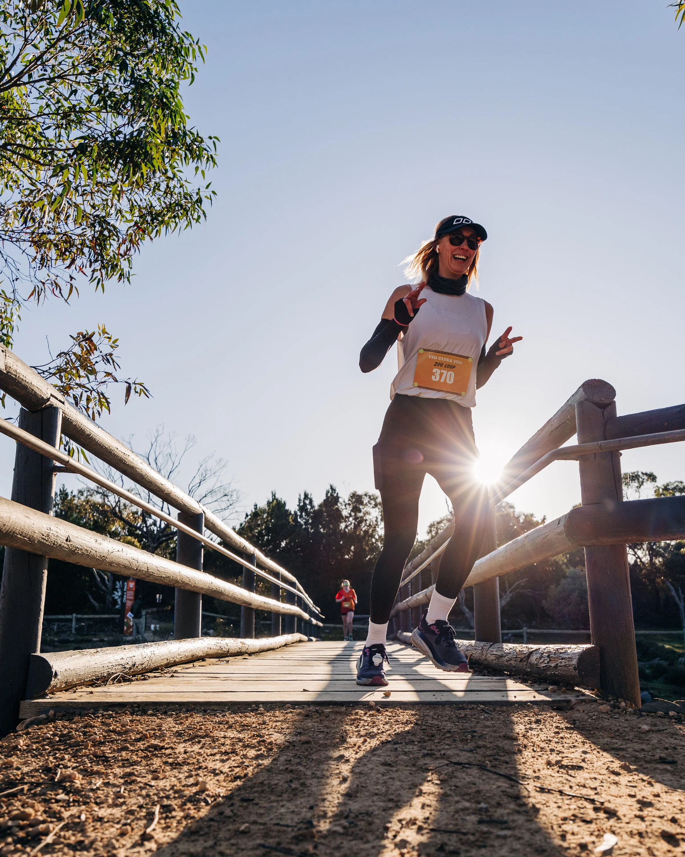 A woman running on a wooden bridge during a race, wearing sunglasses, a race bib, and athletic clothing, with the sun shining behind her and other runners in the background.