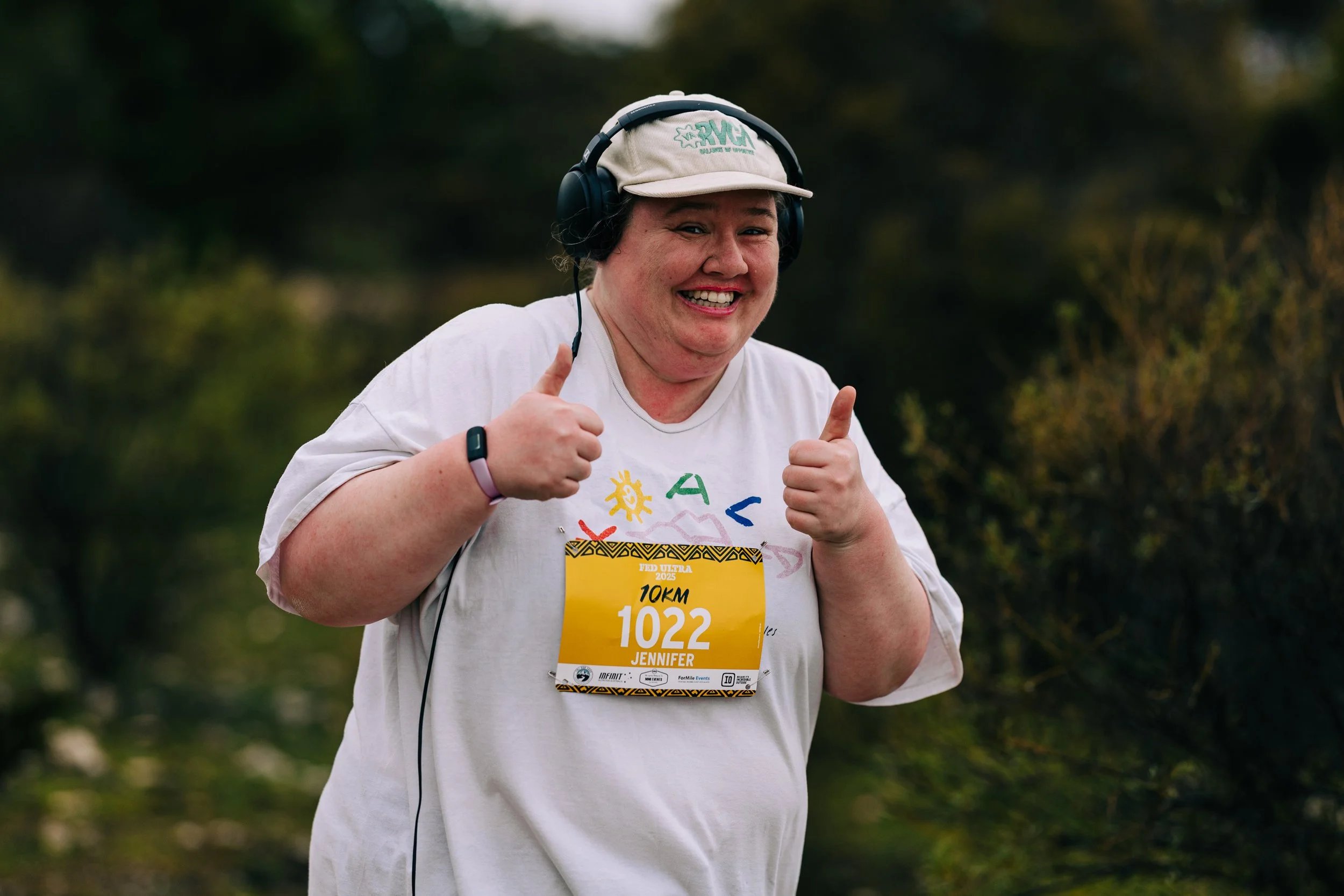 A smiling woman participating in a 10K race, wearing a beige cap, white t-shirt, and headphones, with a bib that reads "Jennifer" and gives a thumbs-up gesture.