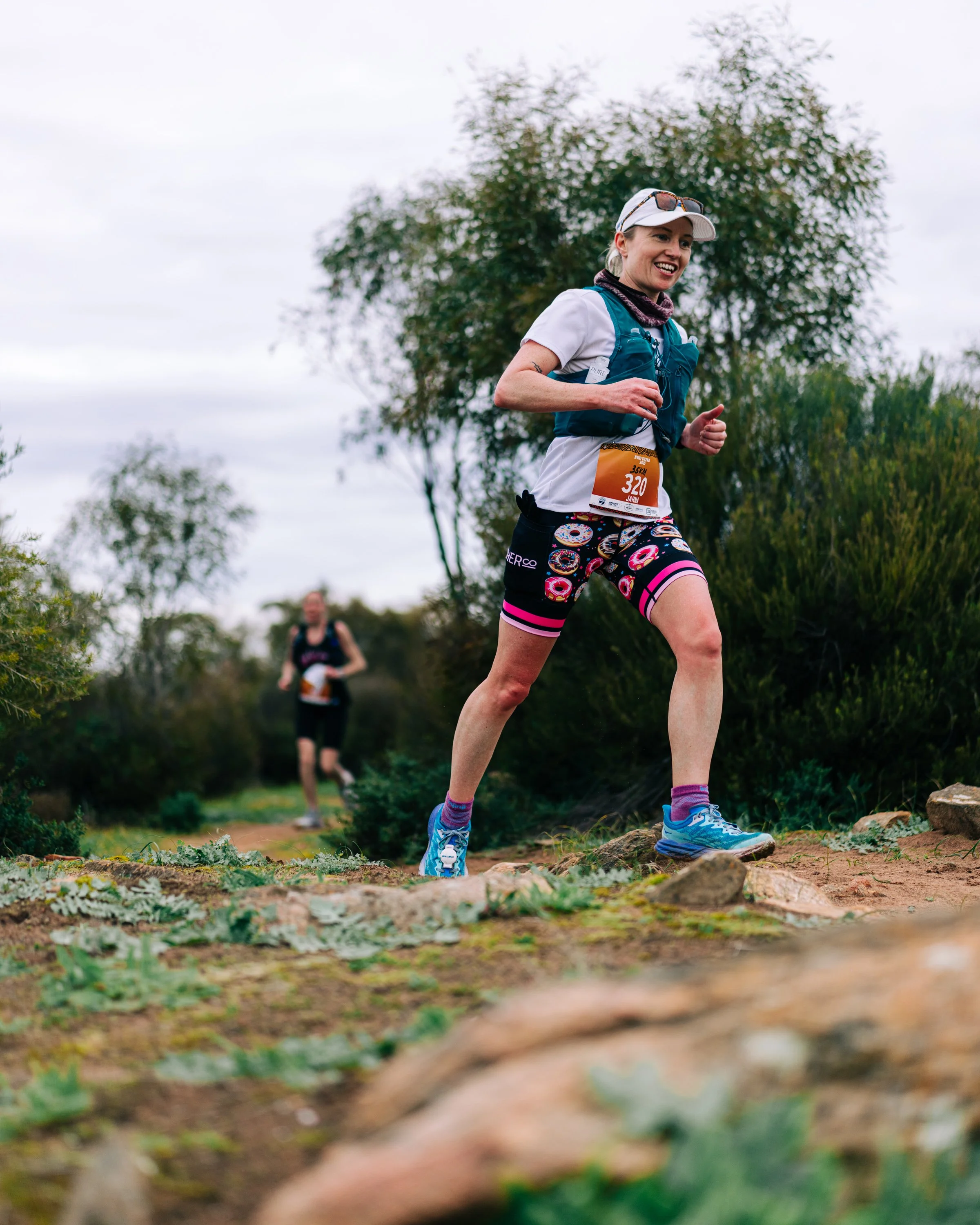 Woman trail running outdoors, wearing a white cap, white shirt, patterned shorts, and running shoes, with another runner in the background among trees and bushes on an overcast day.