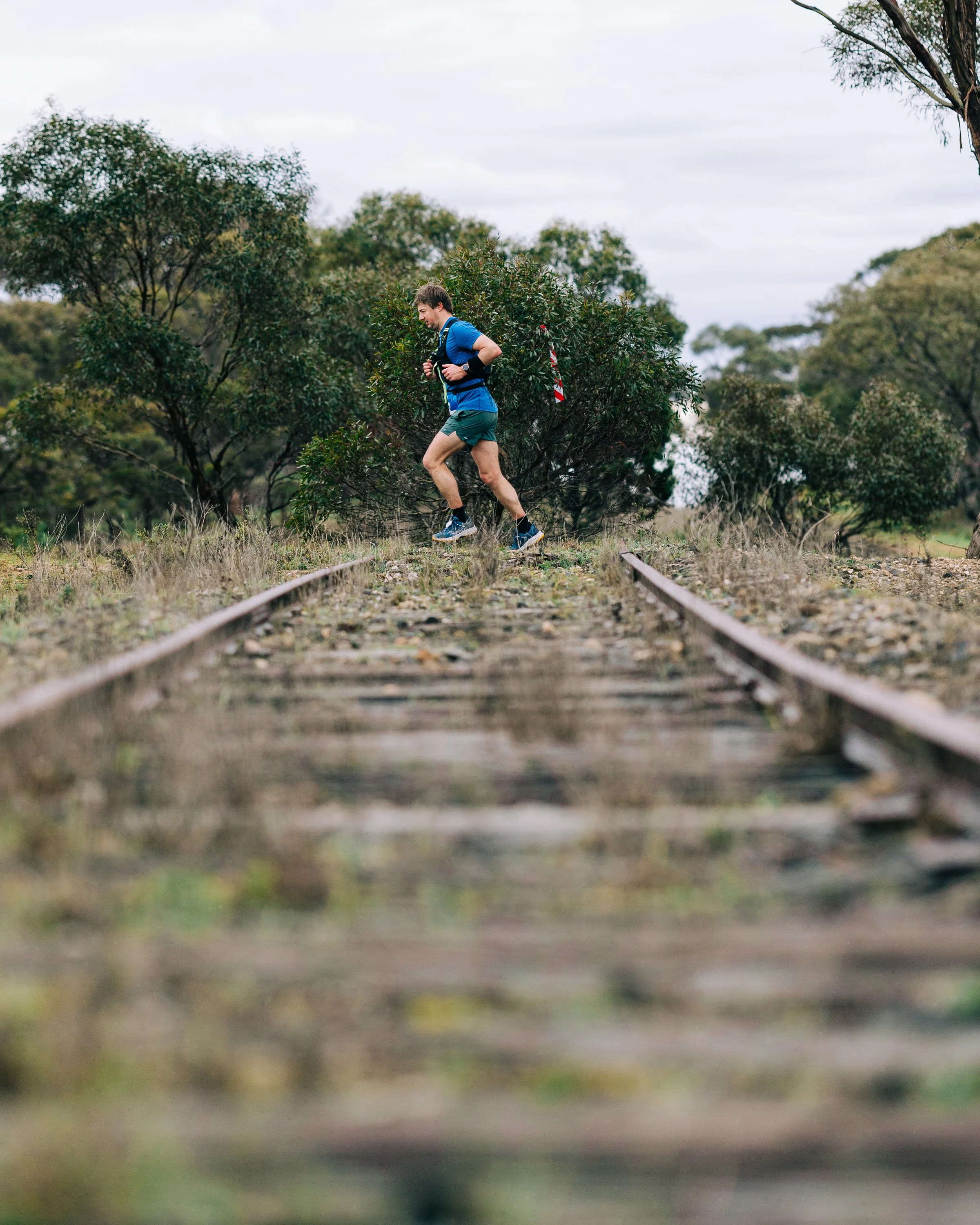 A man running on abandoned train tracks in a rural outdoor setting with trees and overcast skies.