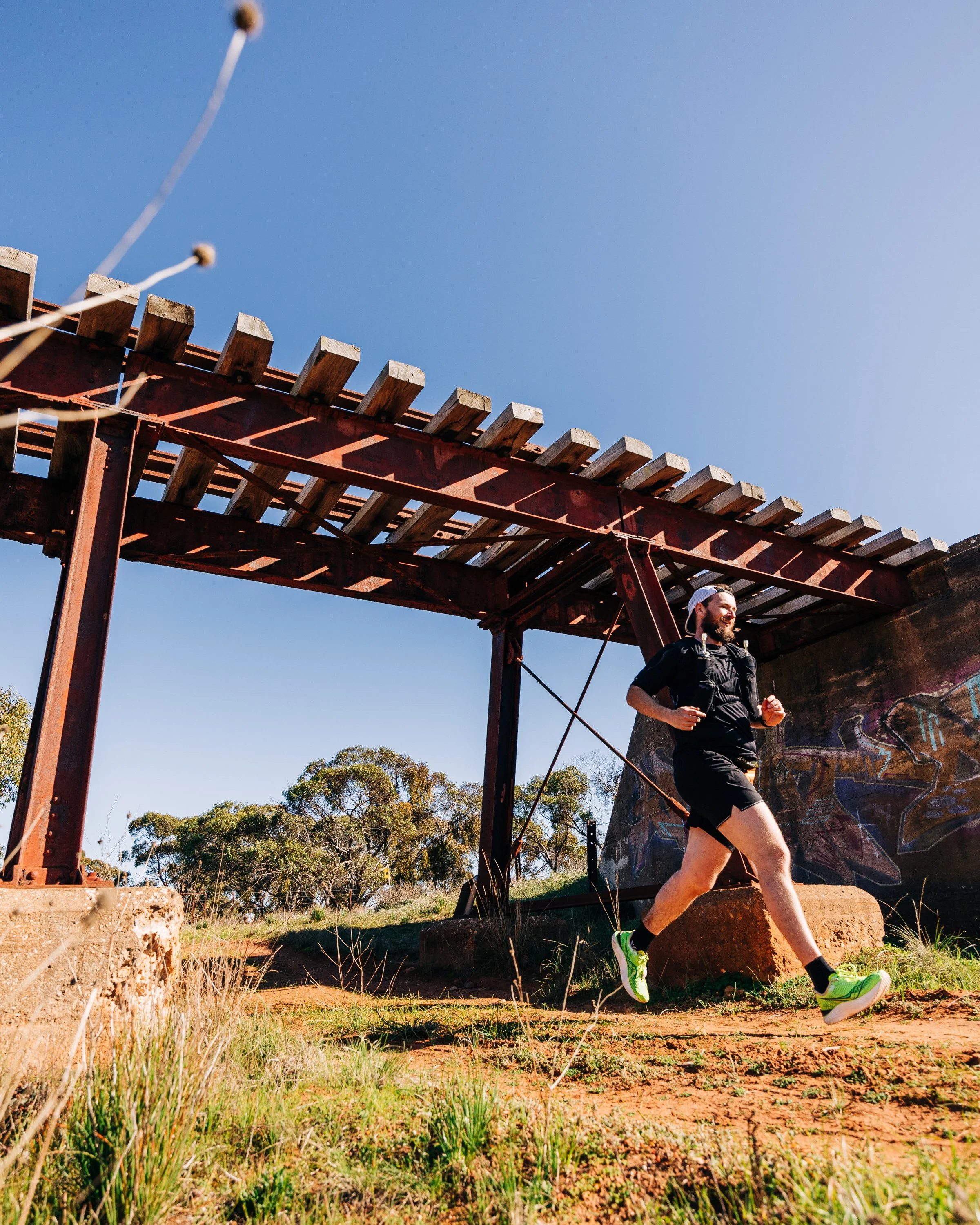 A man running outdoors under a wooden bridge structure with graffiti on the wall, bright blue sky, and trees in the background.
