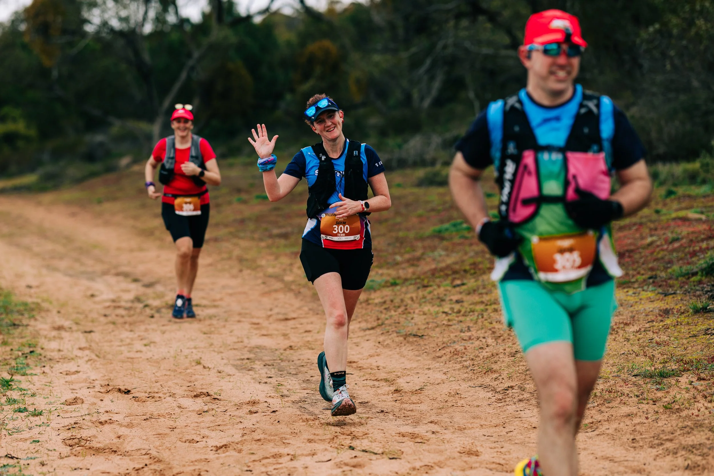 Three runners on a dirt trail in a forest, smiling and waving, wearing colorful running gear and race bibs.