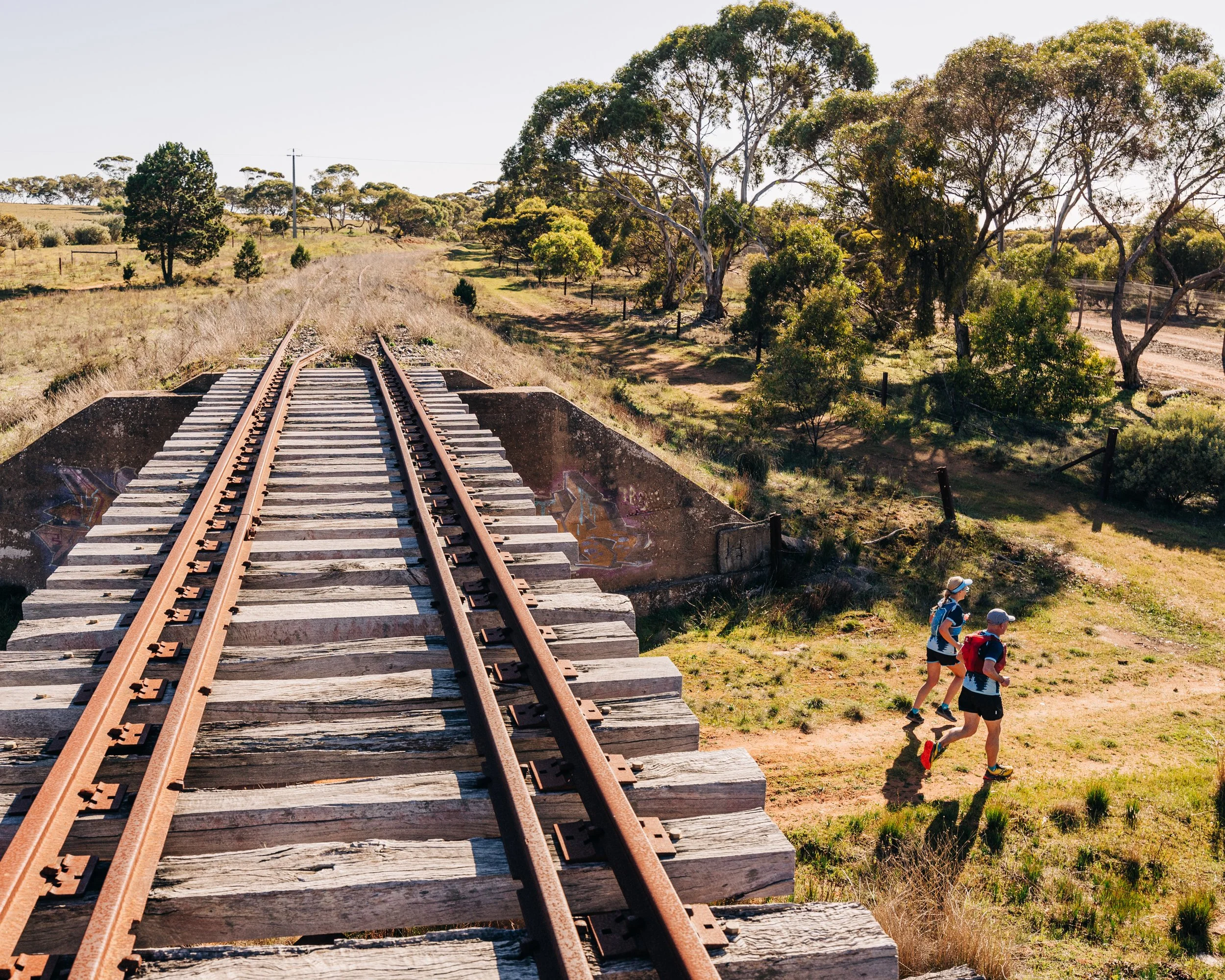 Two hikers walking on a dirt trail near a railway bridge in a grassy, tree-filled landscape on a sunny day.