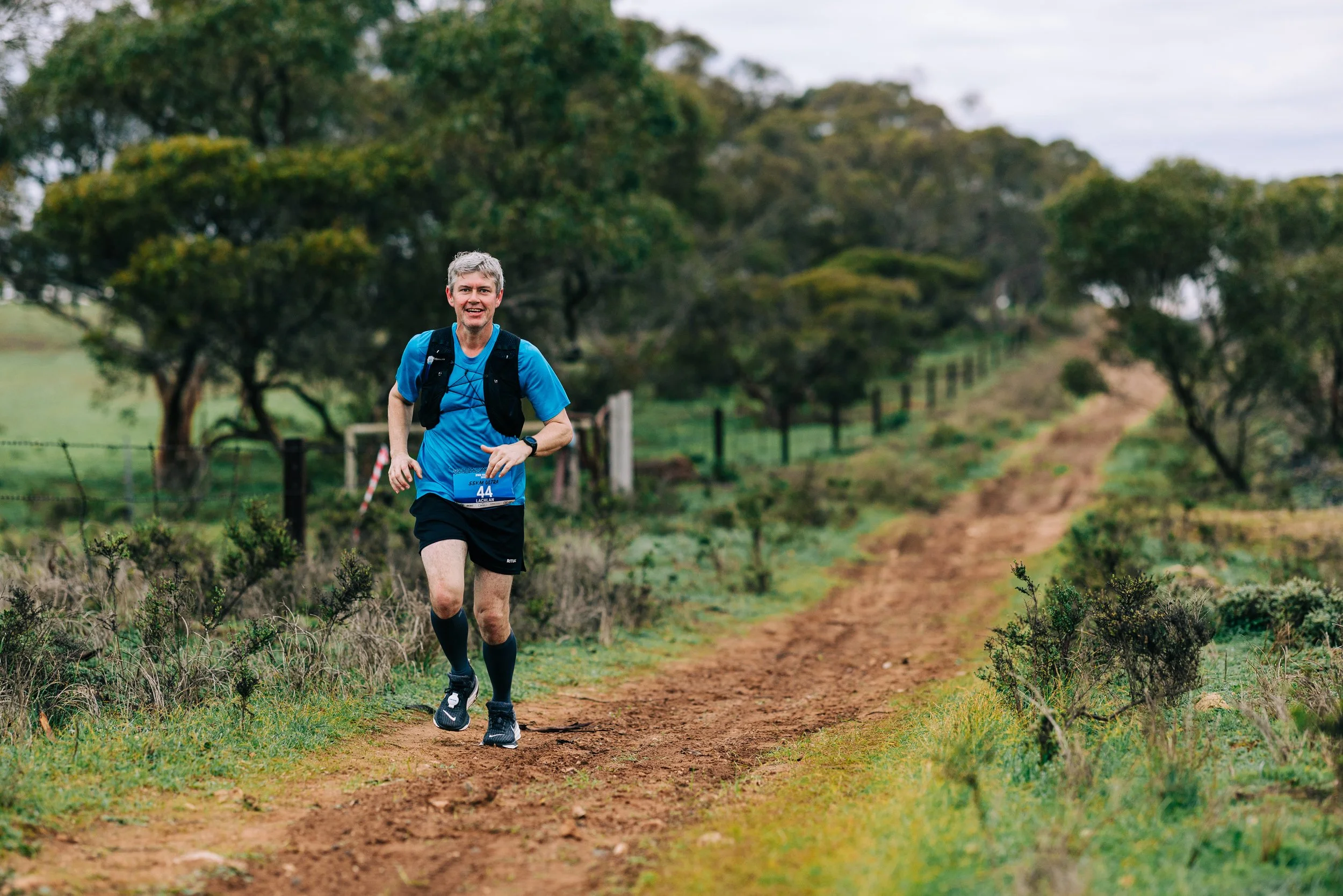 A man in a blue shirt running on a dirt trail in a green rural area with trees and bushes.