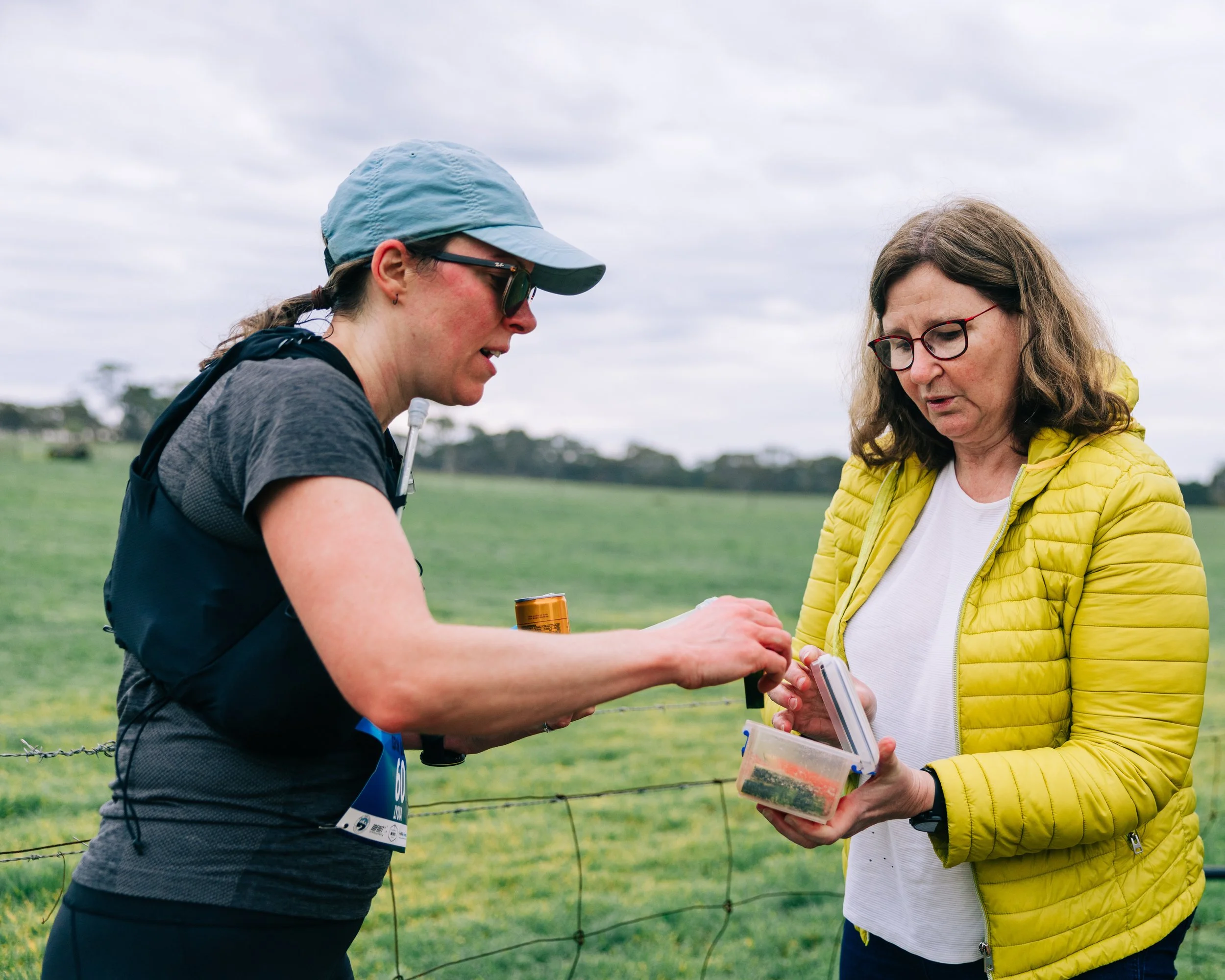 Two women outdoors next to a fence, one holding a small container and the other handing her something, in a green field under a cloudy sky.