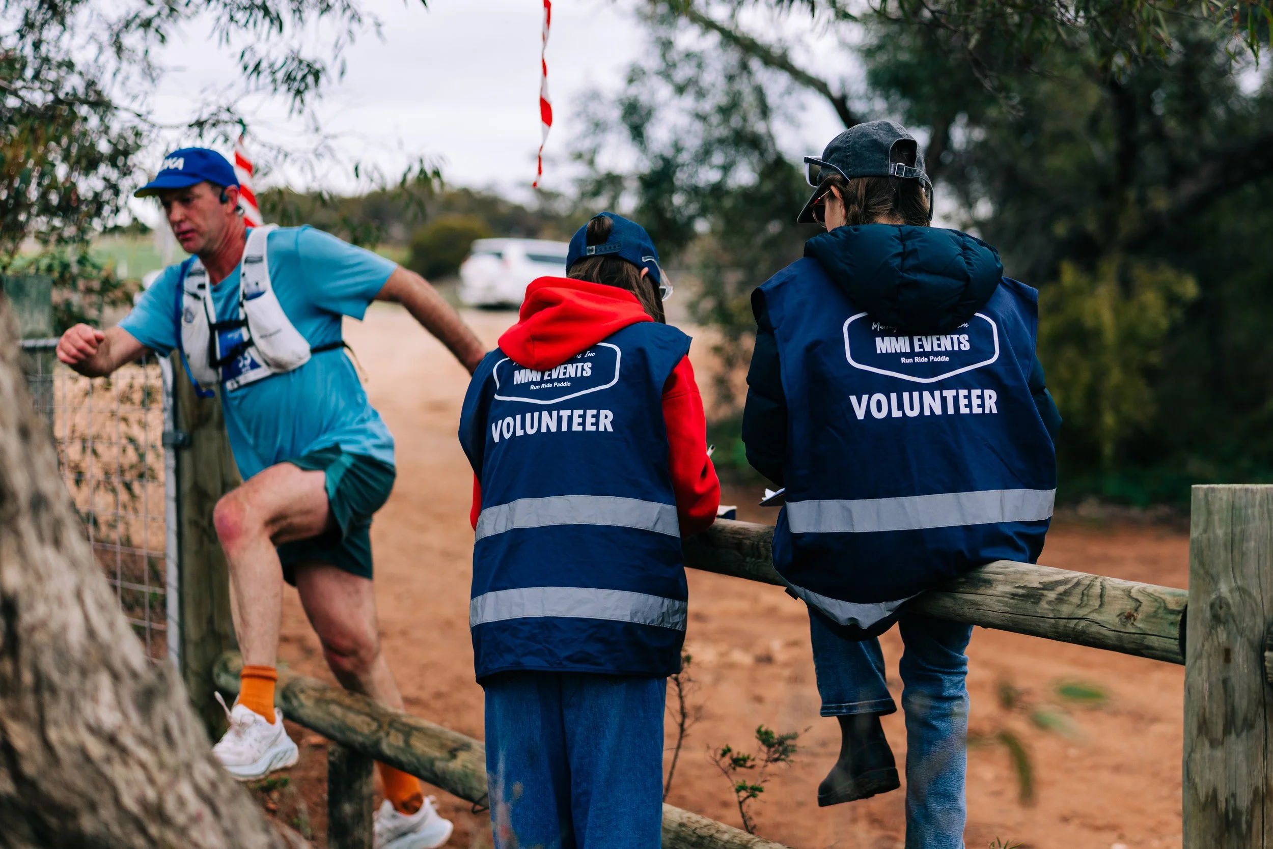 Two volunteers sitting on a wooden fence talking, and a man in athletic gear running past them during an outdoor running event.