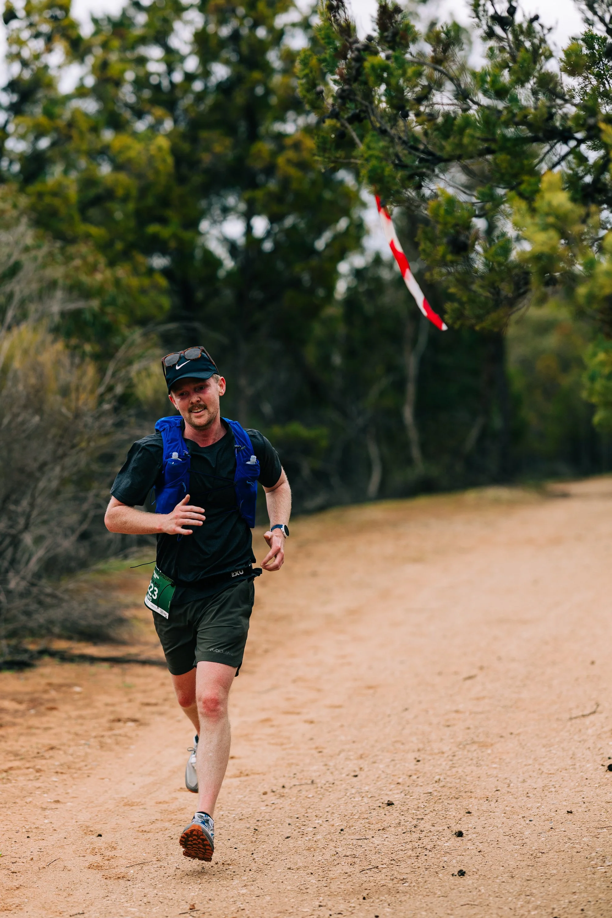 A man is running on a dirt trail through a wooded area, wearing athletic clothing and a race bib, with a determined look on his face.