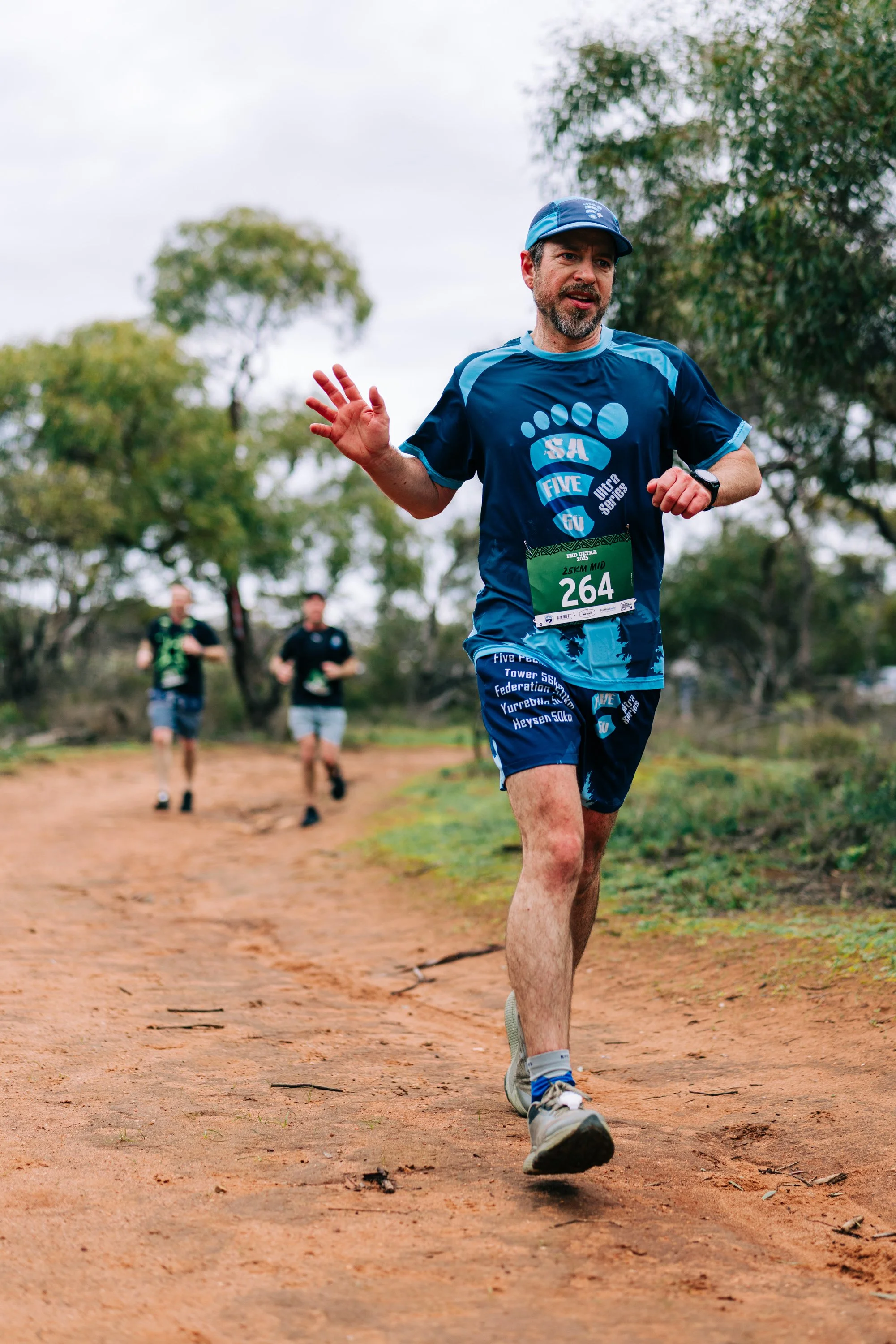 A man running on a dirt trail during a race, wearing a blue athletic outfit, a race bib with the number 264, and a cap, with two other runners behind him and trees in the background.