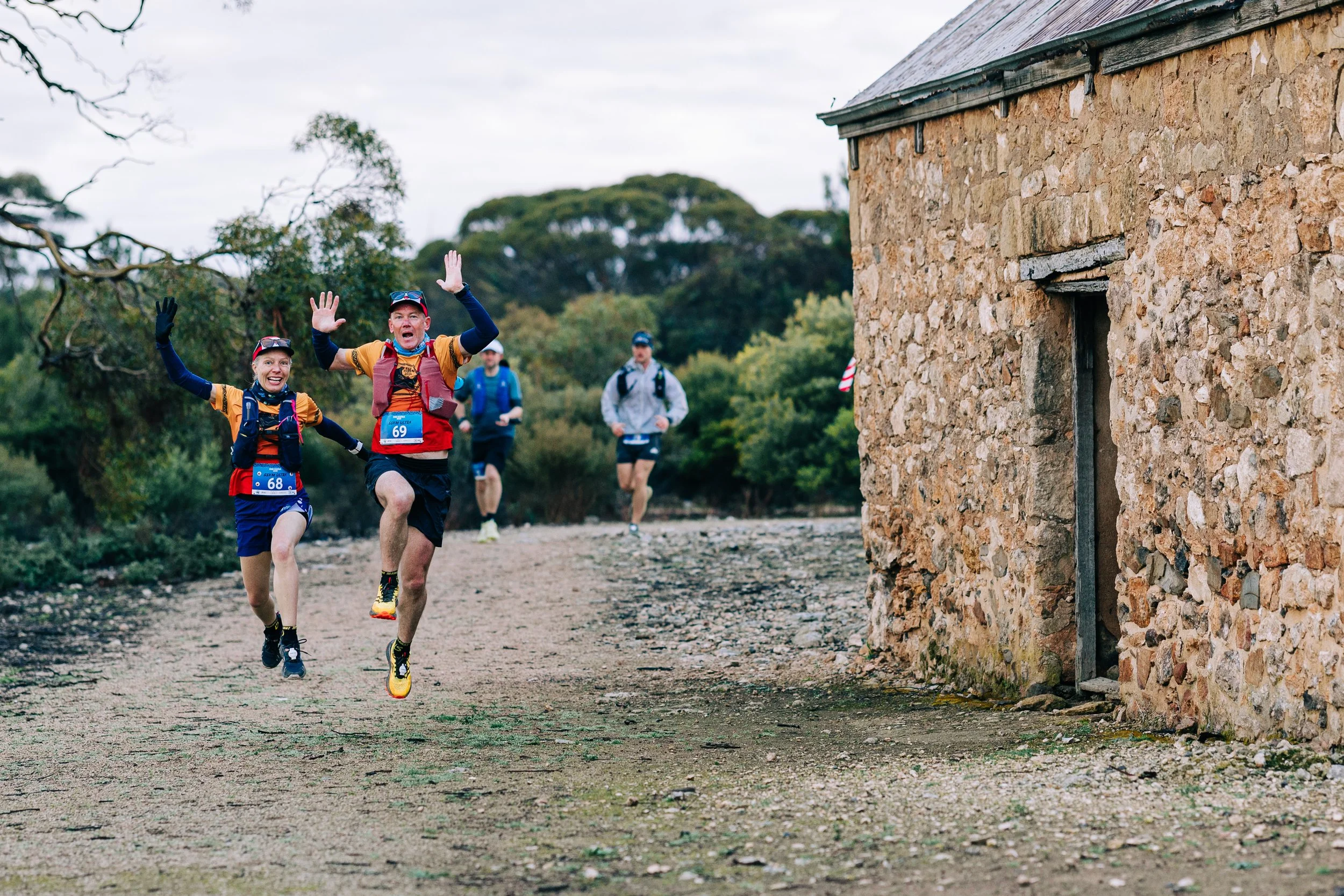 Four runners participating in a trail race, two of them are in the foreground smiling and waving at the camera, while two others are behind them on a gravel trail with trees and an old stone building on the side.