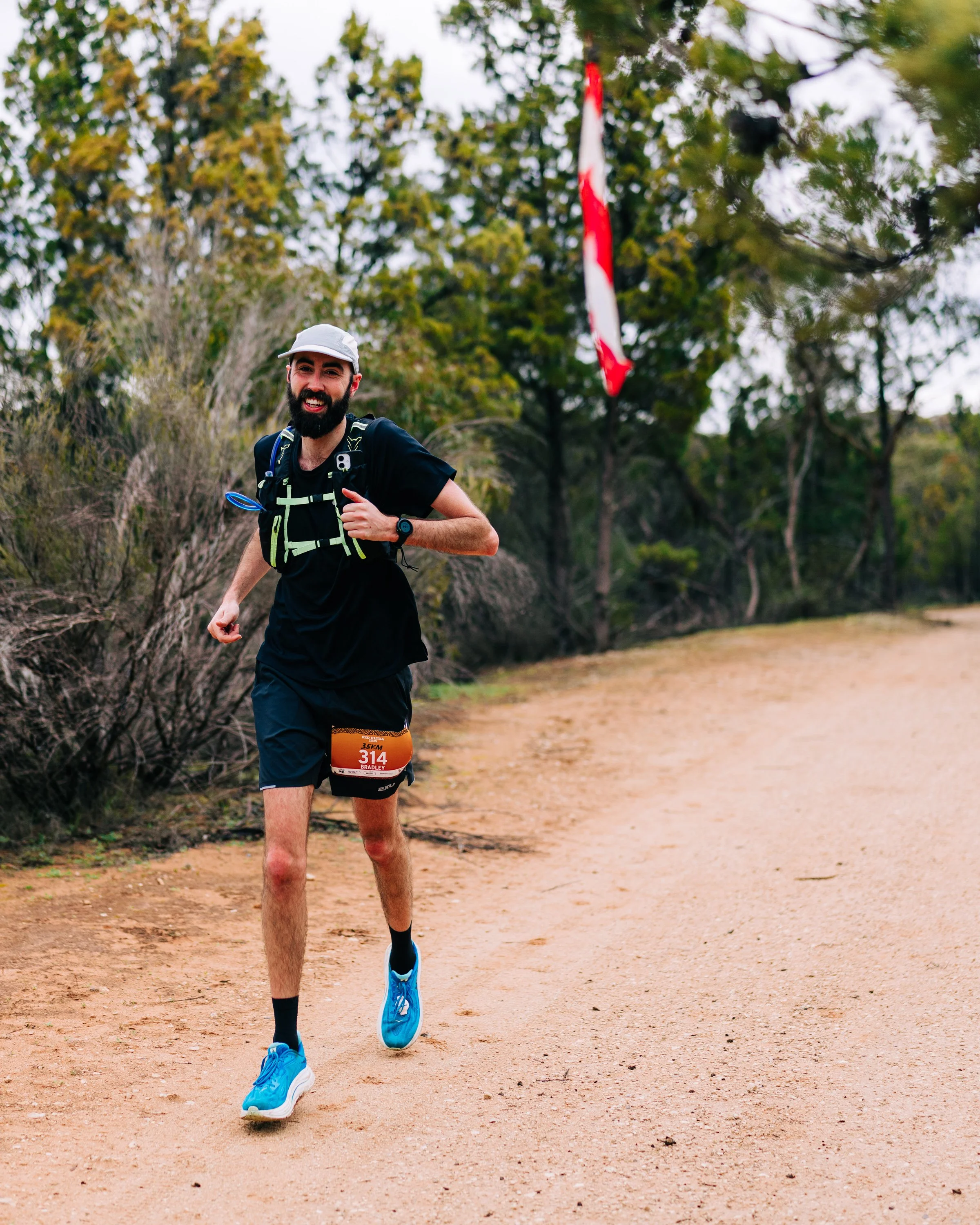 A male trail runner with a beard and white cap joyfully running on a dirt trail through a forested area during a race, wearing a race bib numbered 314, a black t-shirt, black shorts, bright blue running shoes, and a hydration vest.