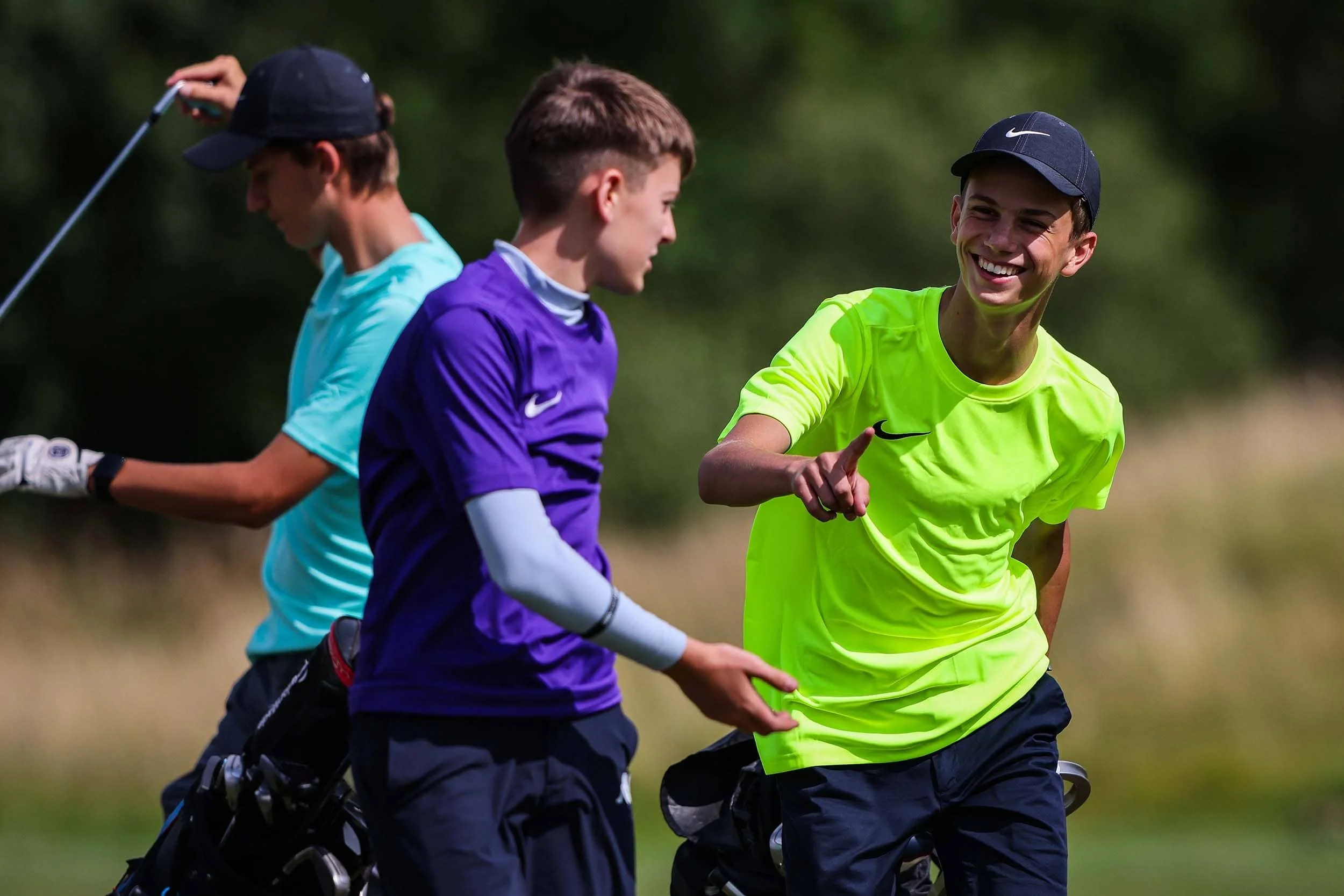 Tres jóvenes golfistas en un campo de golf, uno con una camiseta verde neón brillante, sonriendo y señalando, el segundo con una camiseta morada y blanca, hablando, y el tercero con una camiseta azul claro y un palo de golf, practicando o jugando al golf.