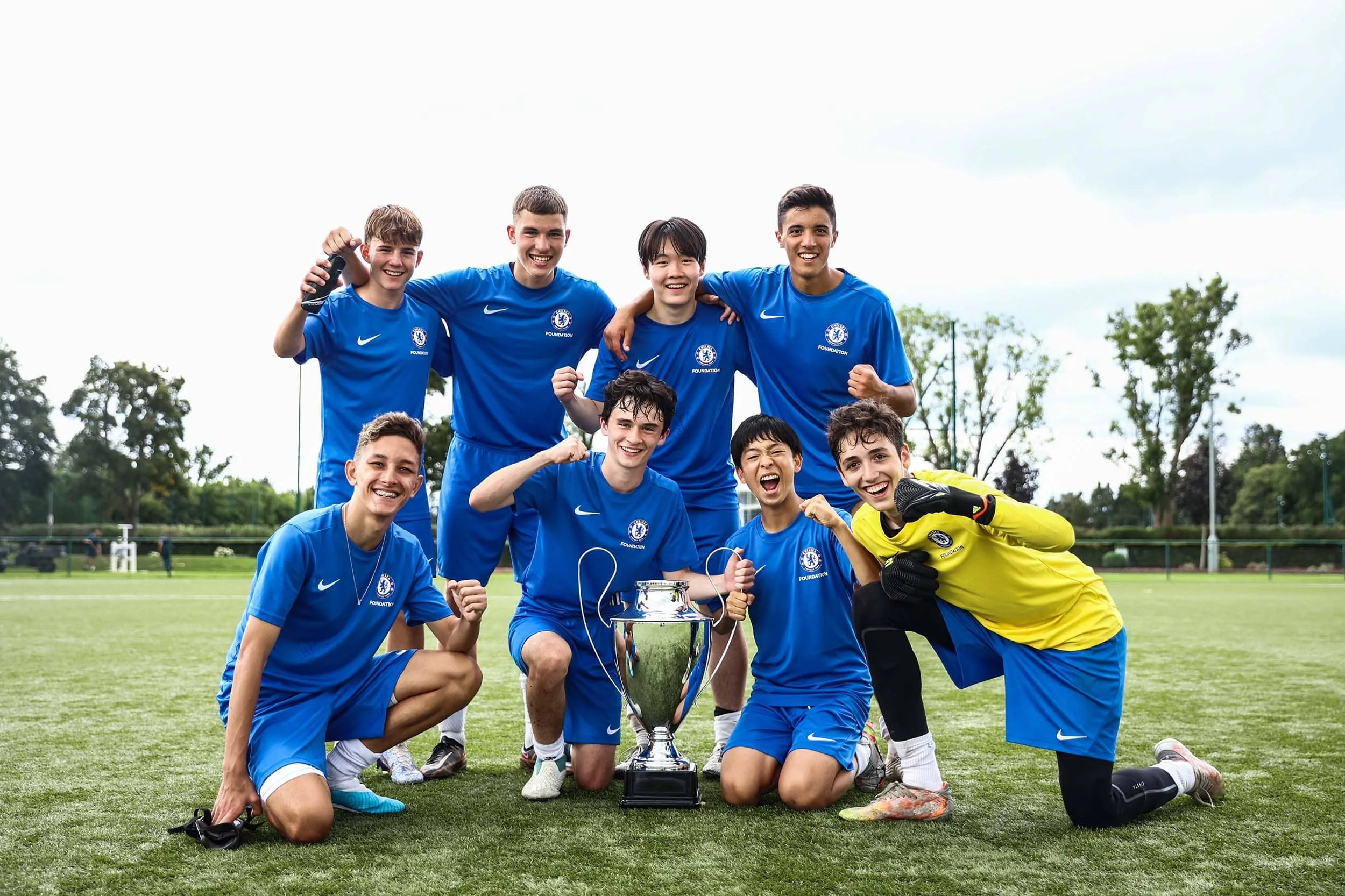 Group of young soccer players in blue jerseys celebrating on the field with a trophy, some kneeling and some standing, outdoors on a grassy field with trees in the background.