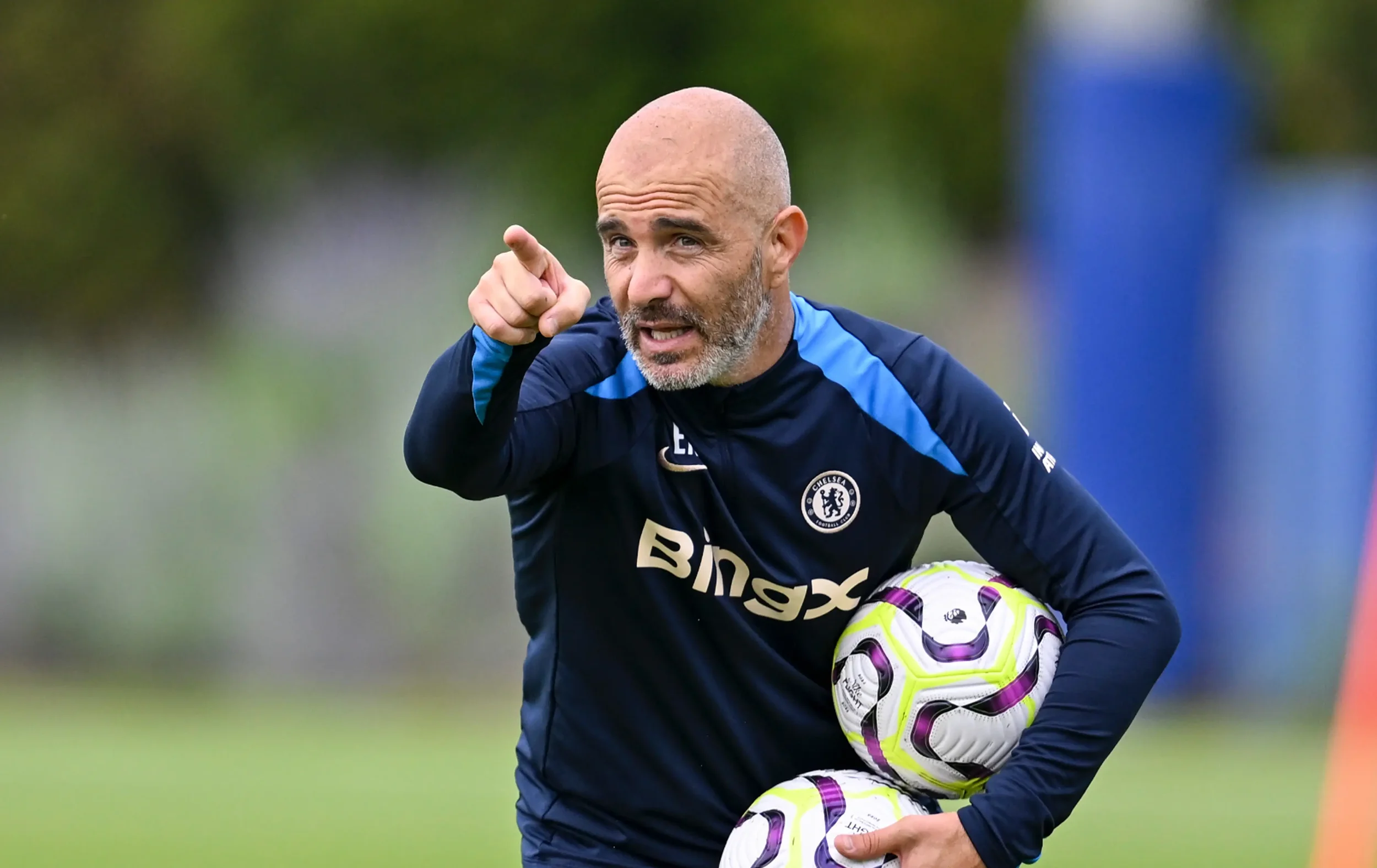 A man in a Chelsea soccer training jersey holds three soccer balls while pointing and speaking during practice on a grassy field.