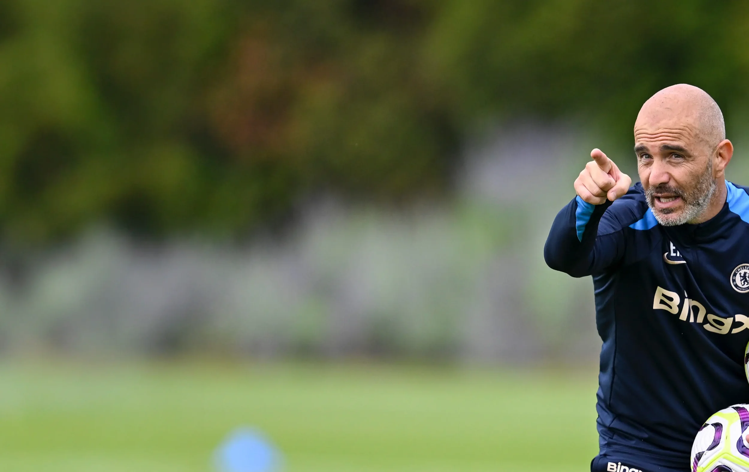 A man in sports attire, bald and with a beard, pointing and speaking during a sports practice or game outdoors, holding a soccer ball, with a blurred natural background.
