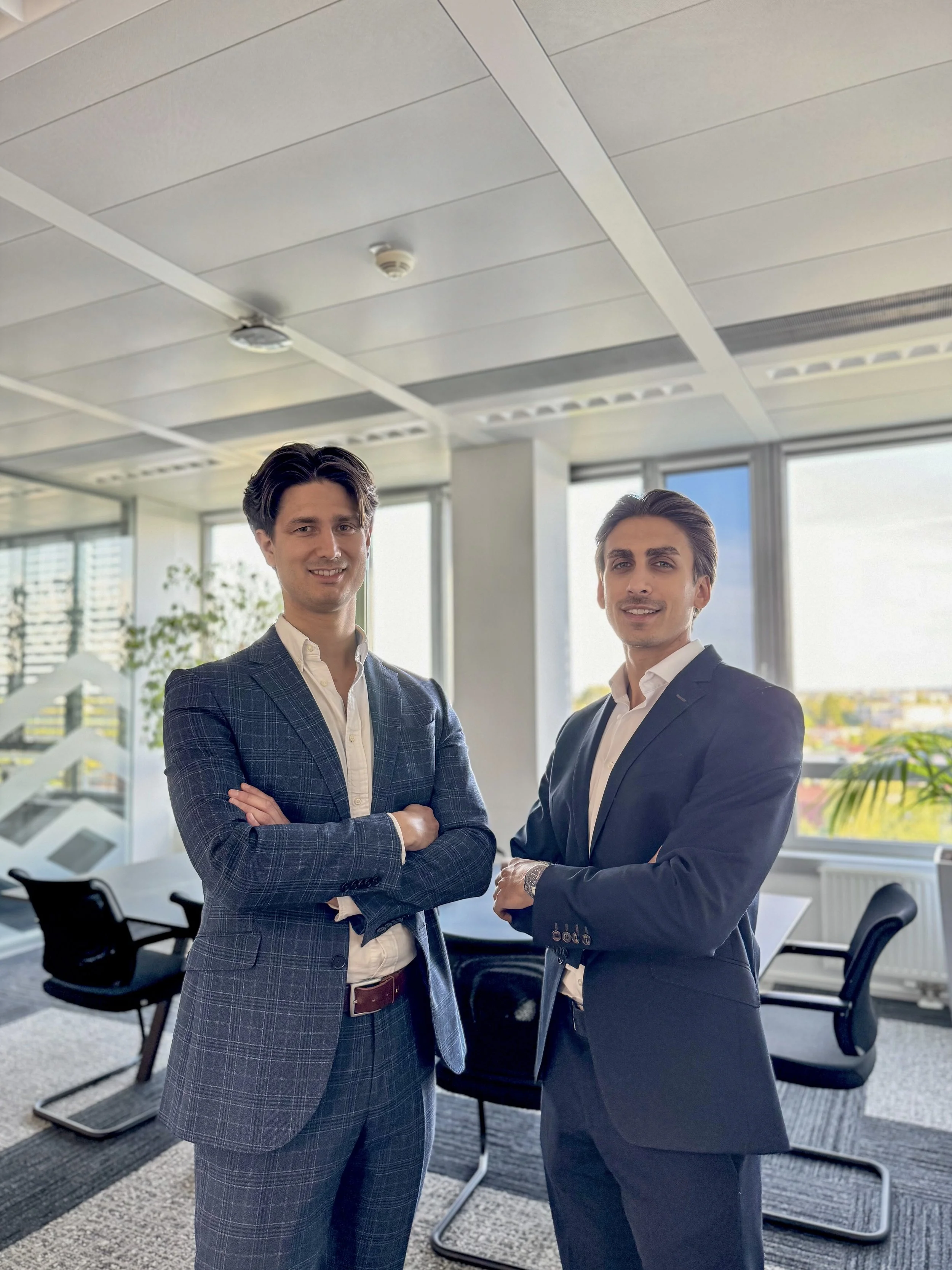 Two men in suits standing in an office with large windows, office chairs, and a conference table.
