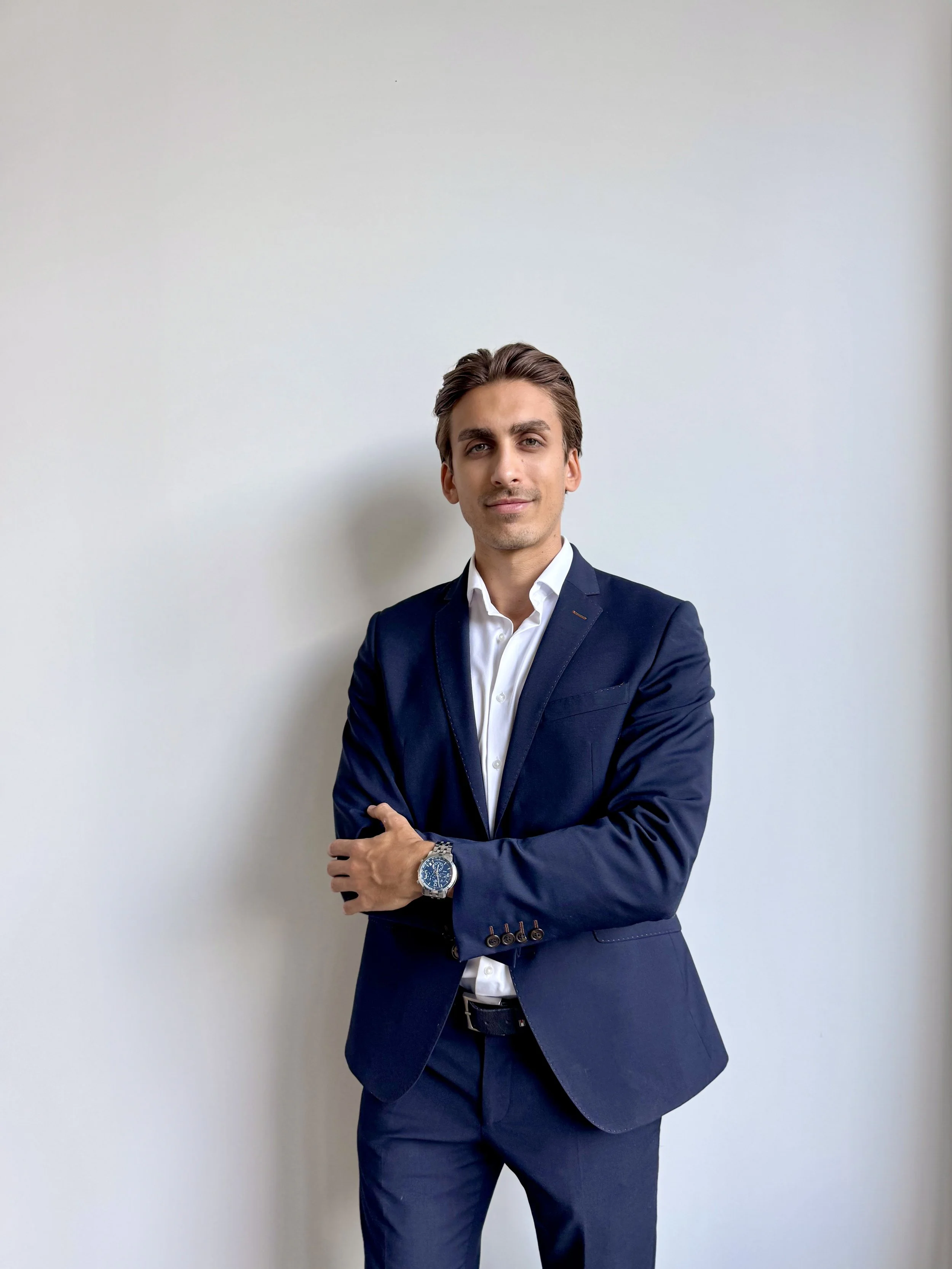 A man in a navy blazer and white shirt standing against a plain white wall.
