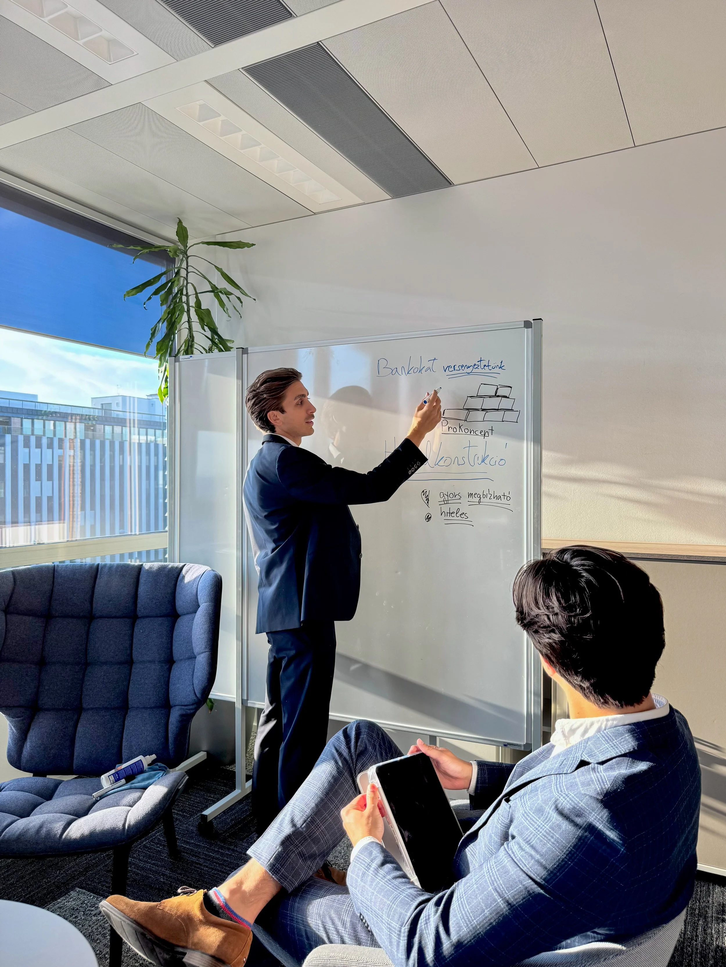 A man in a business suit is writing on a whiteboard in a modern office with large windows. Another man, also in business attire, is sitting and looking at him, holding a tablet. There is a blue armchair nearby and a tall potted plant by the window.