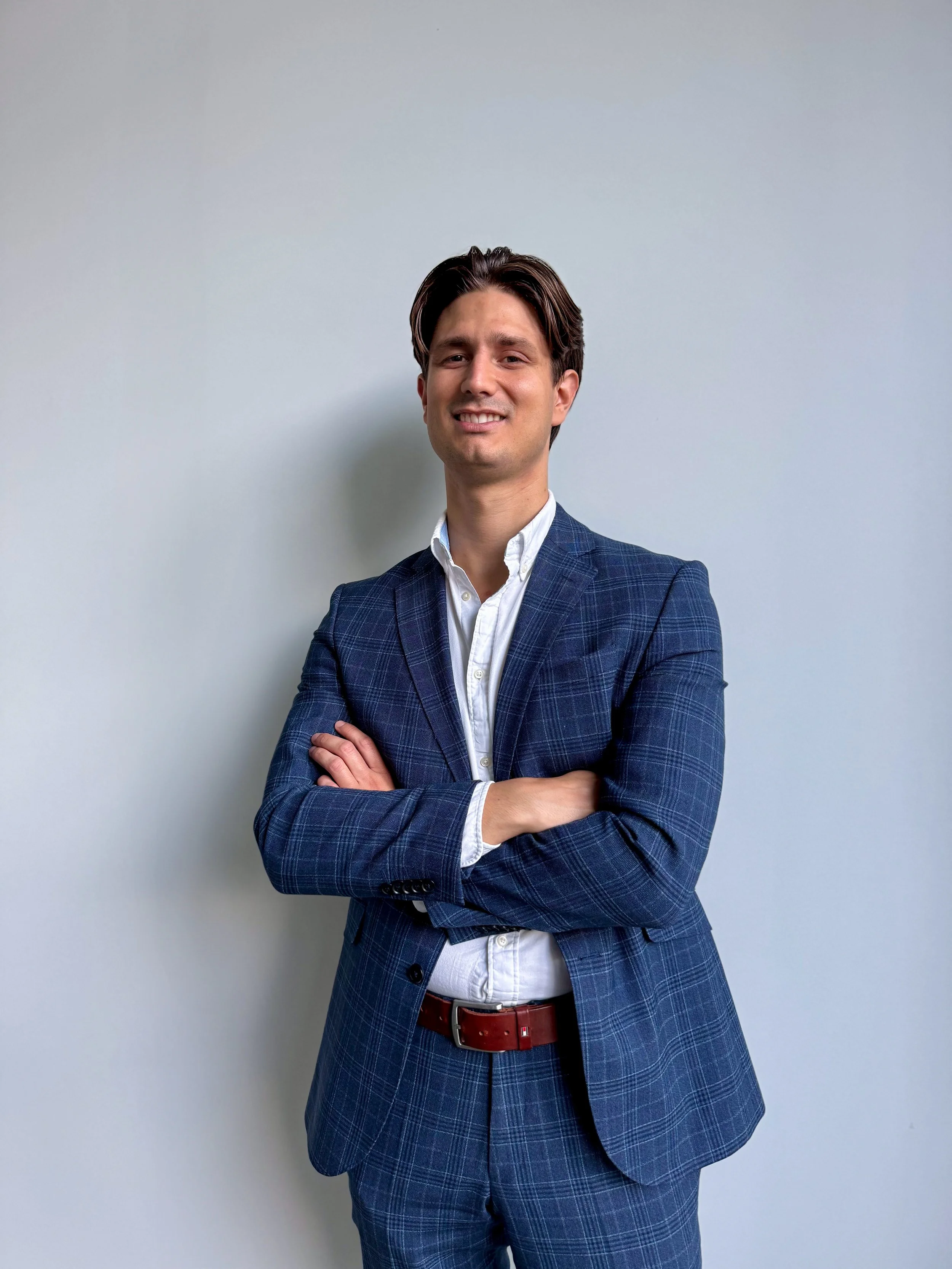 A man in a blue plaid suit with a white shirt, standing with arms crossed, smiling, in front of a plain white wall.