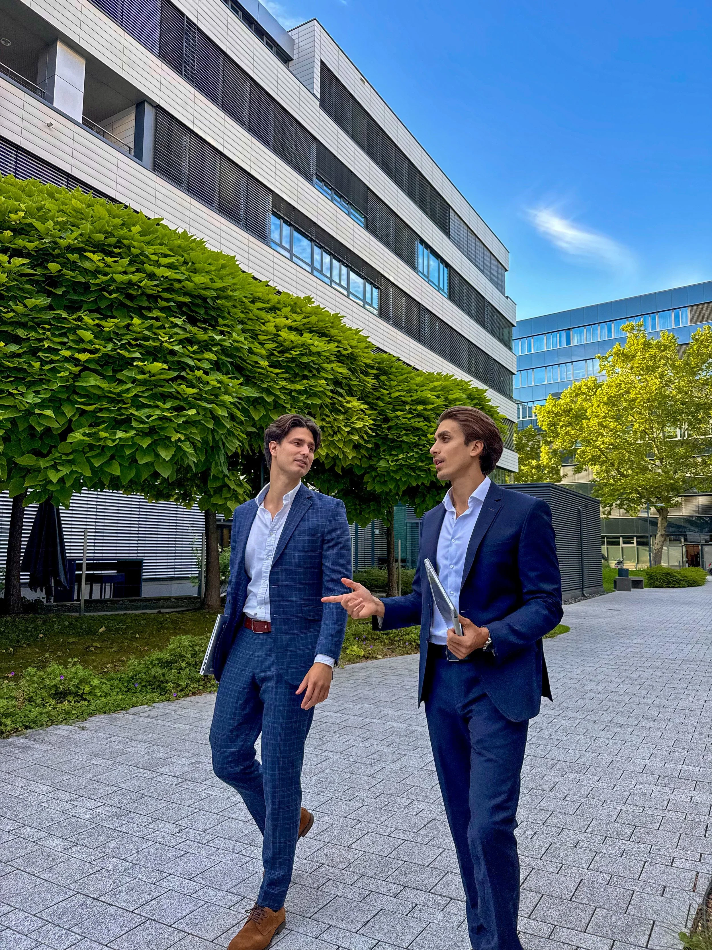 Two men in business suits having a conversation outdoors in an urban setting with modern office buildings, trees, and a clear blue sky.