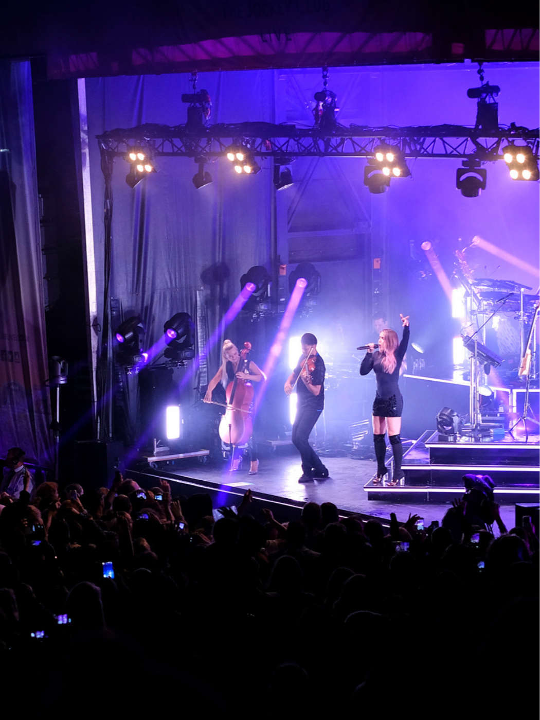 Concert stage with Clean Bandit performing, including a singer with a microphone, a cellist, and a violinist, illuminated by colourful stage lights and an audience in the foreground.