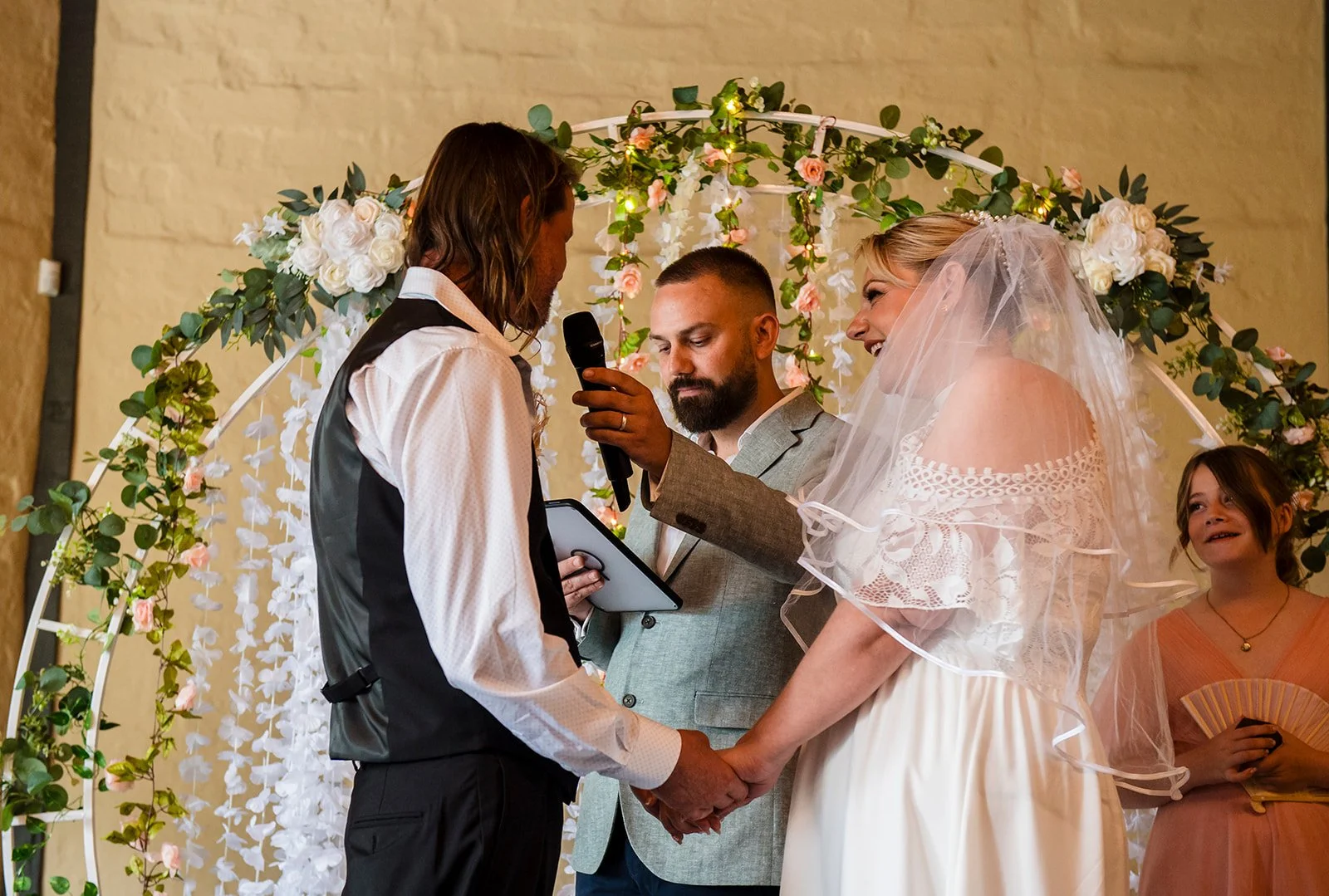 A wedding ceremony with a bride and groom at the altar, surrounded by seated and standing guests in a decorated indoor venue with large plants, wooden walls, and hanging lights.