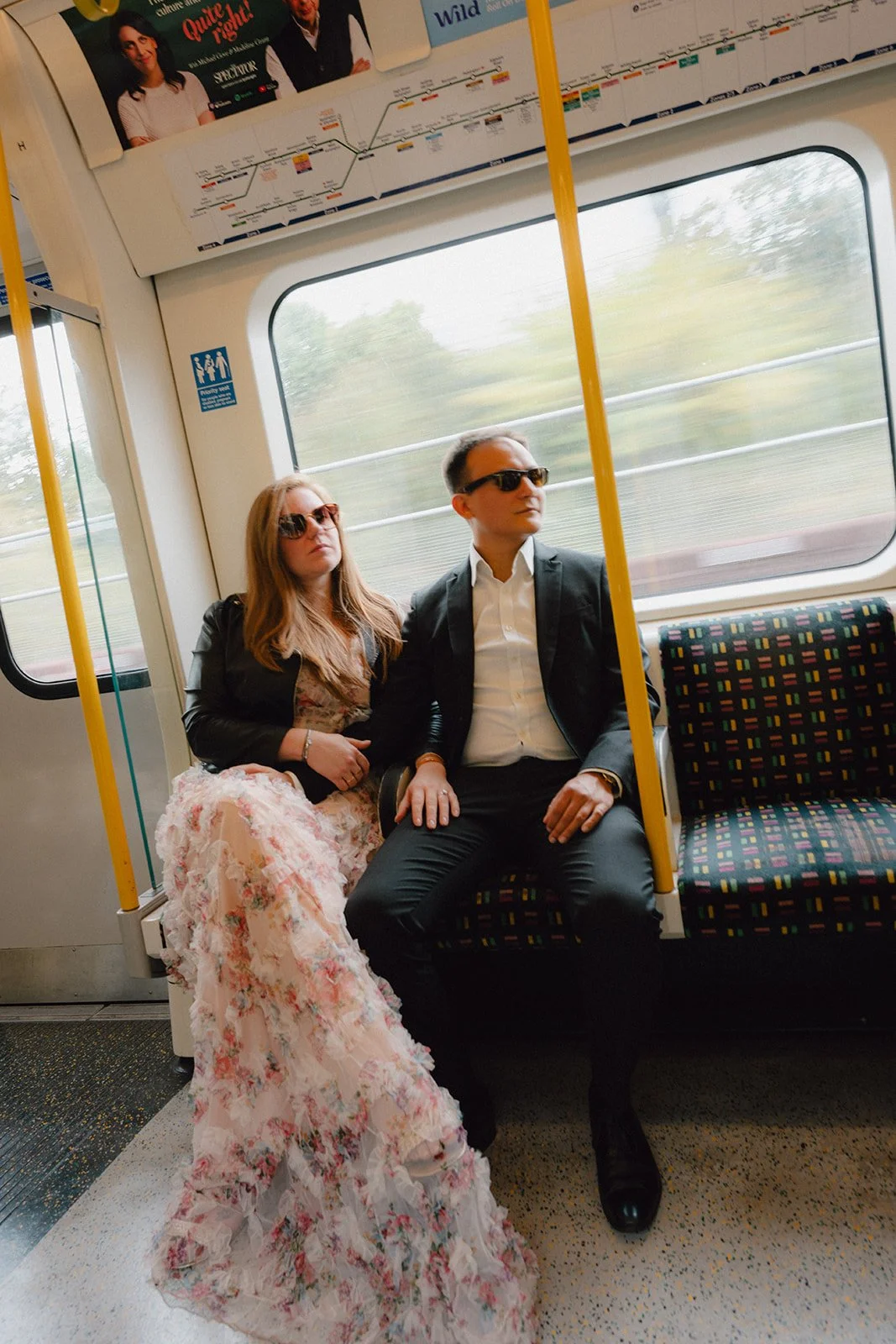 Deux personnes assises dans un train, une femme en robe longue à motifs floraux et un homme en costume noir, tous deux portant des lunettes de soleil.