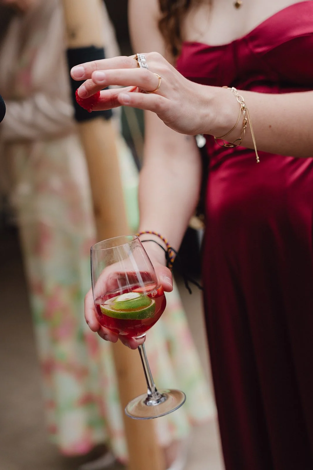 Une femme en robe rouge tient un verre à vin rouge avec des tranches de citron vert. Elle porte des bijoux, dont une bague en or et une chaîne délicate. D'autres personnes en vêtements colorés sont visibles en arrière-plan.
