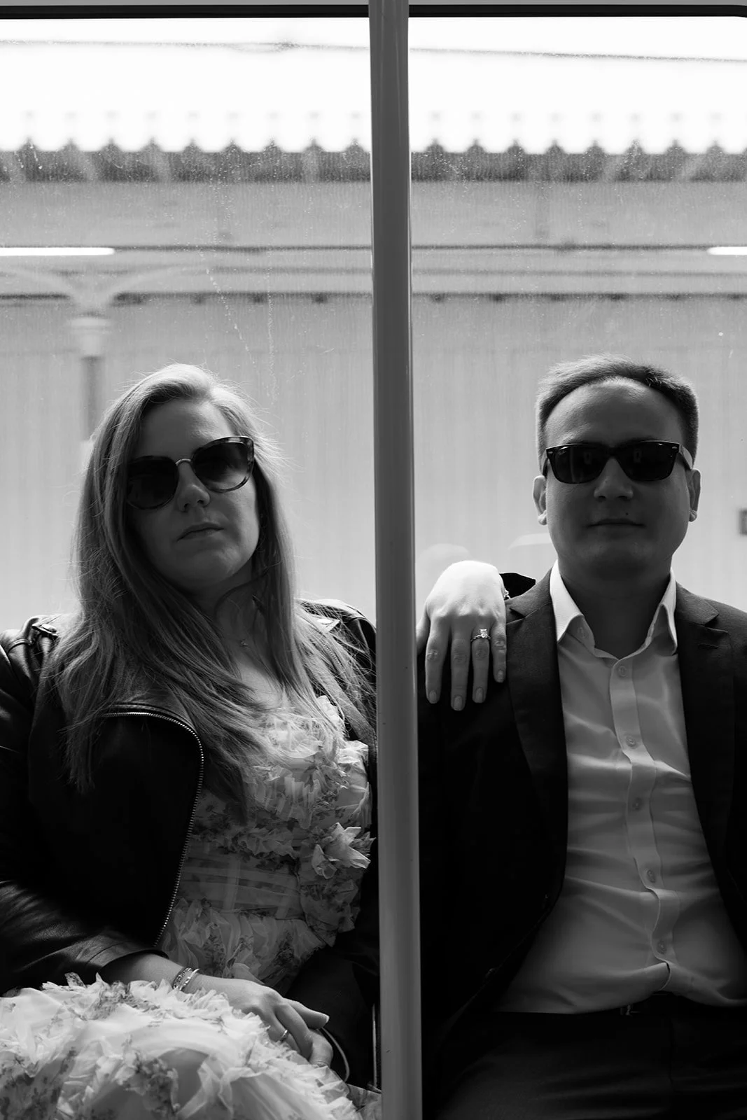 Deux personnes portant des lunettes de soleil, assises dans un train ou un bus, séparées par une barrière centrale, avec une vue floue à l'arrière-plan.