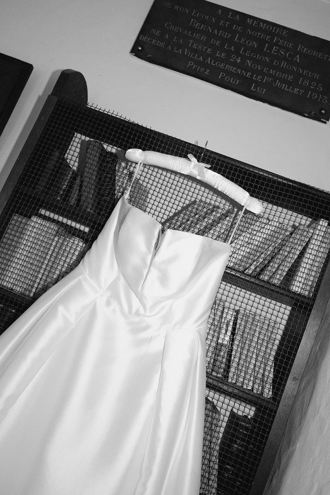 Une robe de mariée blanche suspendue devant une étagère avec plusieurs livres, dans une pièce avec un panneau en arrière-plan. Photo prise au flash, style moderne.